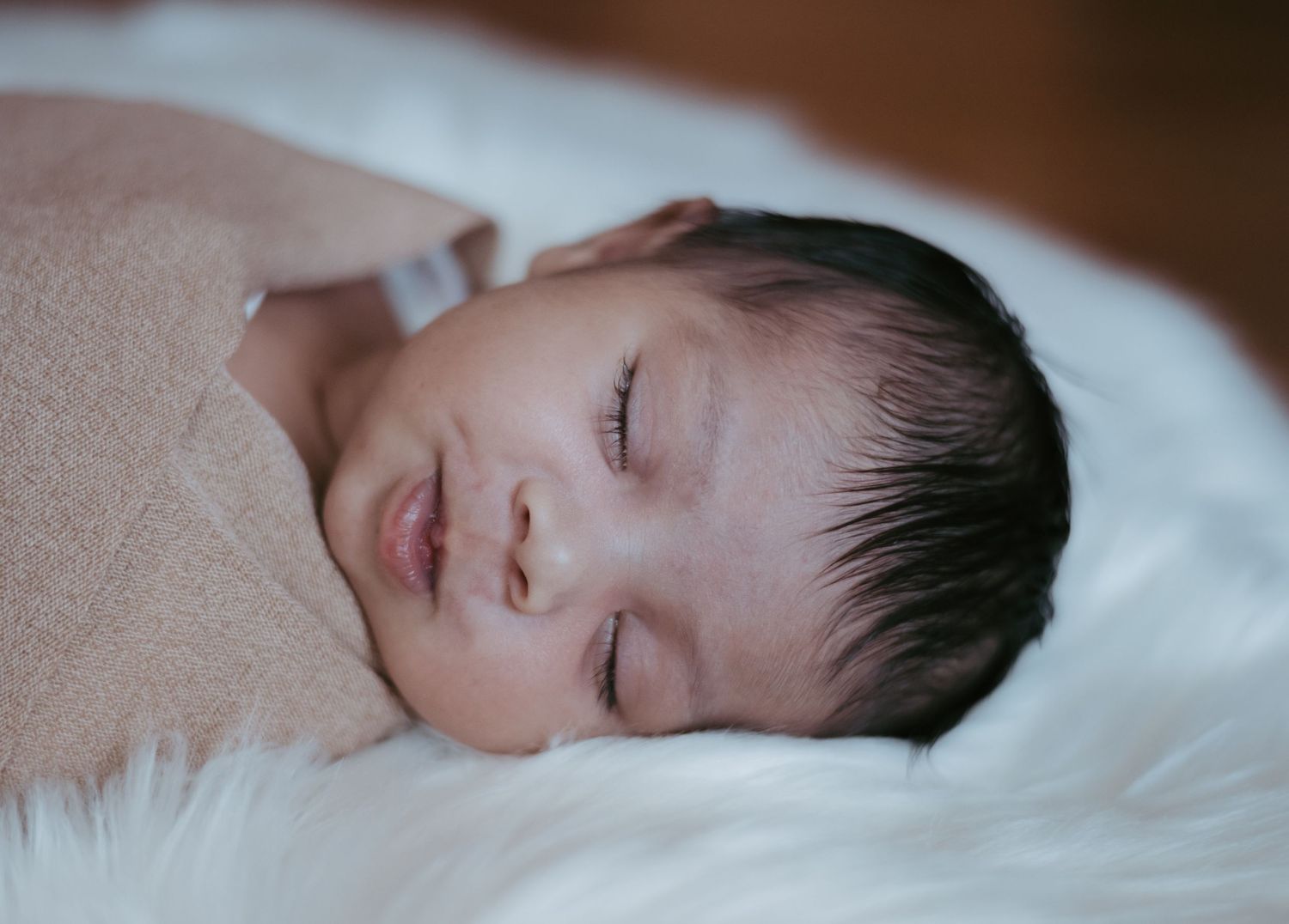Serene sleeping newborn baby with dark hair lying on soft white fluffy surface.