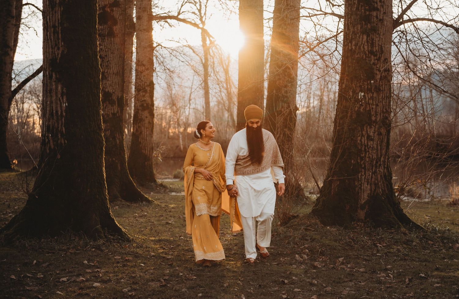 Sikh couple walking hand in hand through golden sunlit forest in traditional Indian wedding attire.