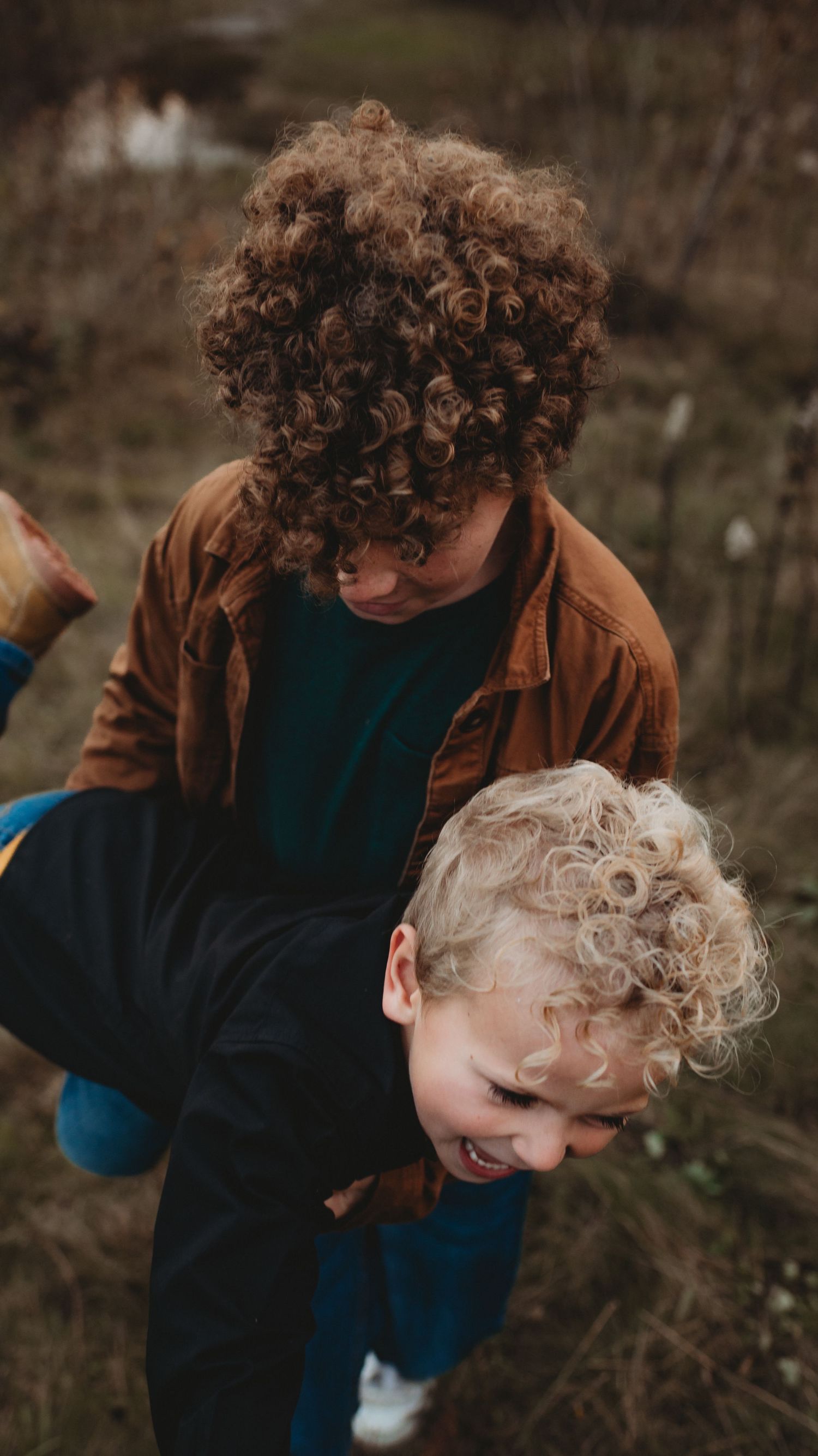 Curly-haired boy playfully lifting a laughing younger blonde boy outdoors in a field.
