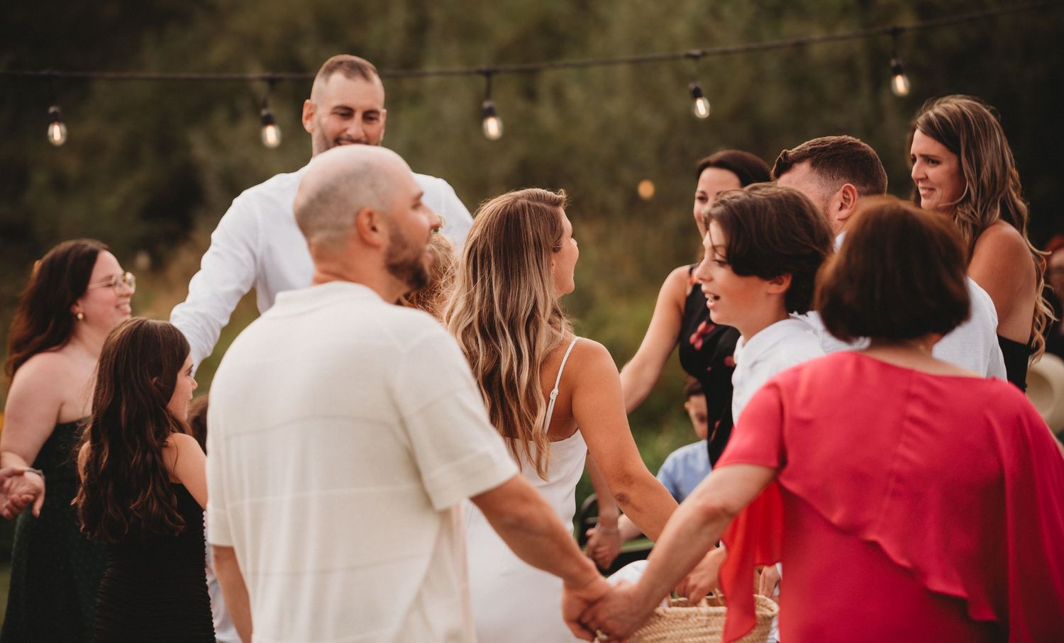 Wedding guests hold hands in a circle at an outdoor evening reception.