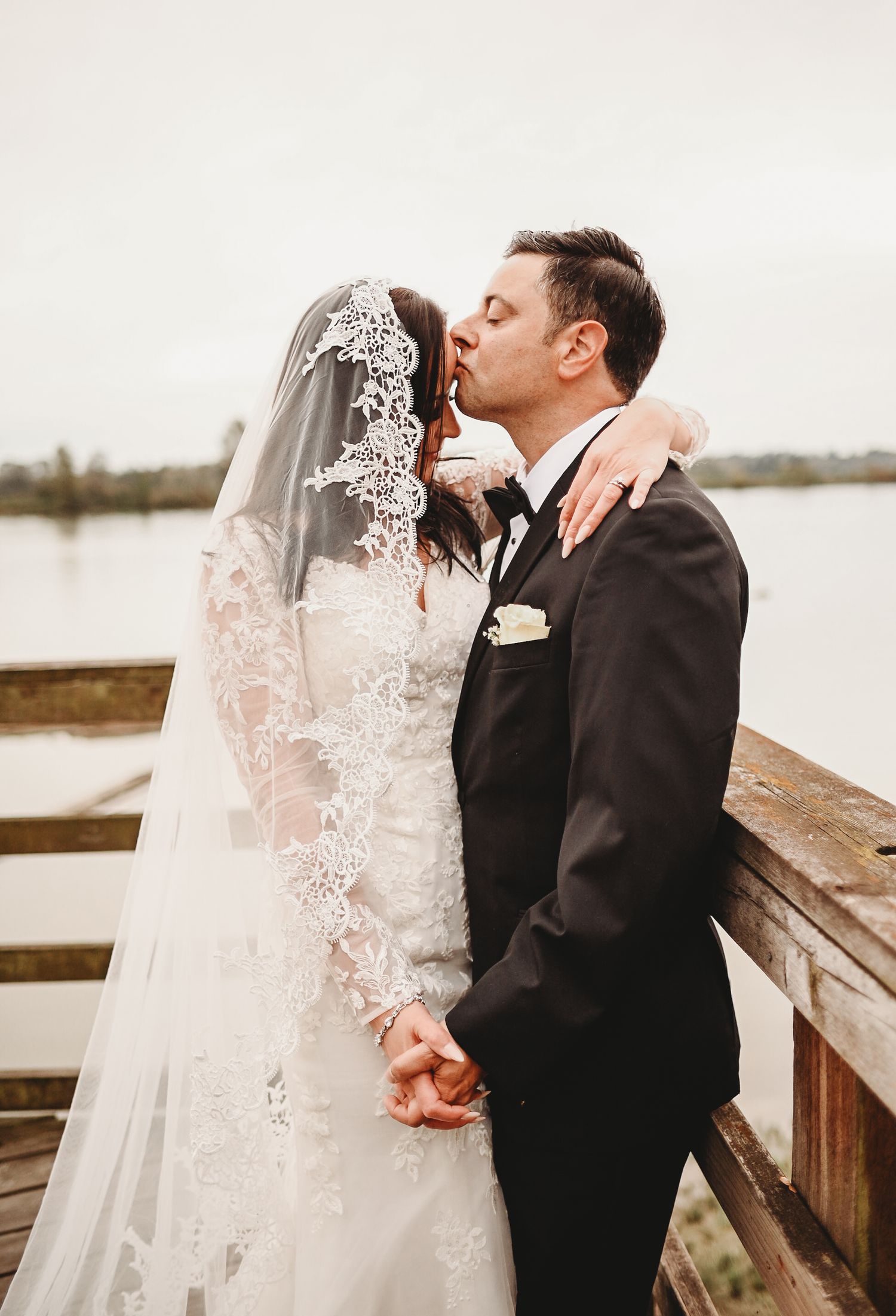 Groom kissing bride on the cheek on a lakeside dock, bride in a lace wedding dress with a lace-trimmed veil.