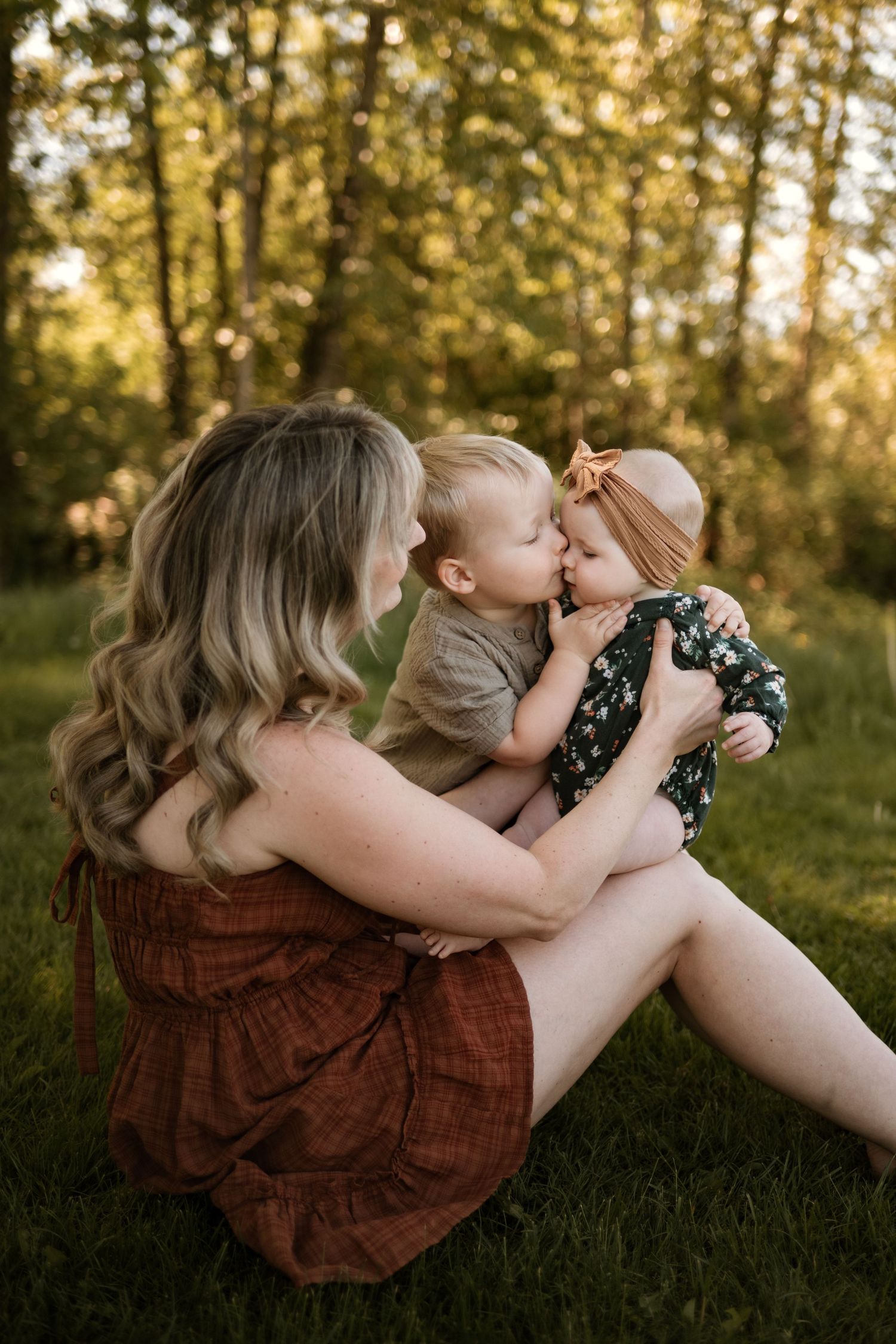Mother sitting on grass holding infant while toddler gives baby a kiss in sunny park.