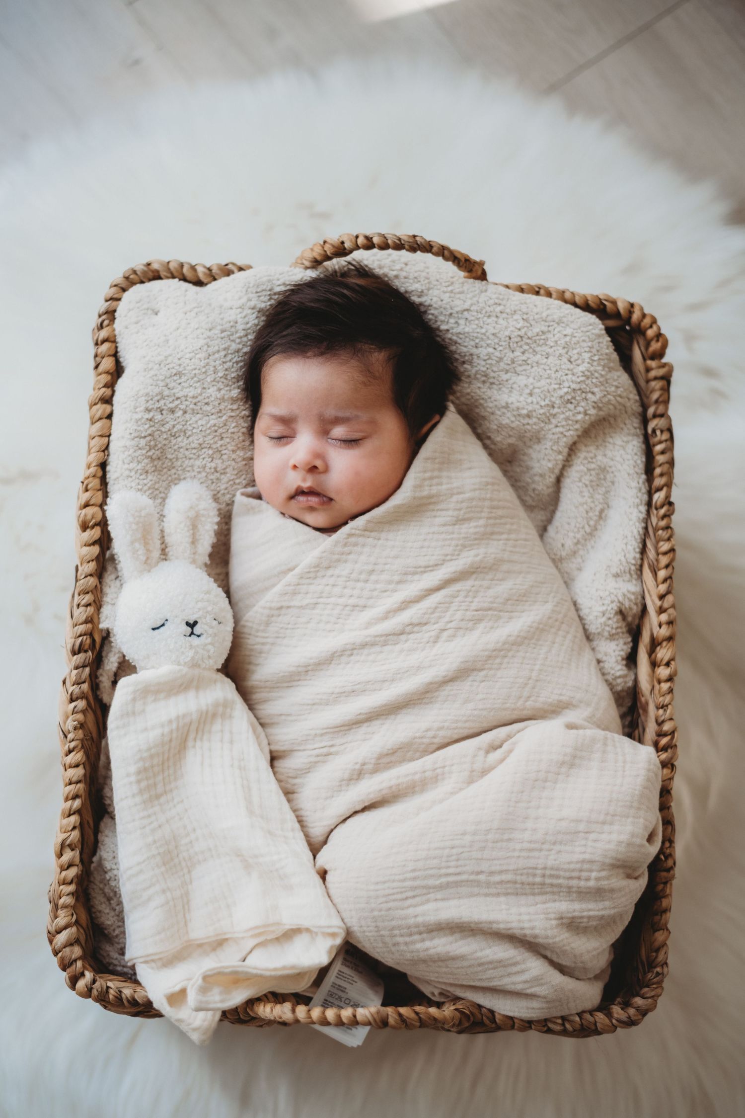 Sleeping newborn baby swaddled in cream wrap with bunny lovey toy in wicker basket.