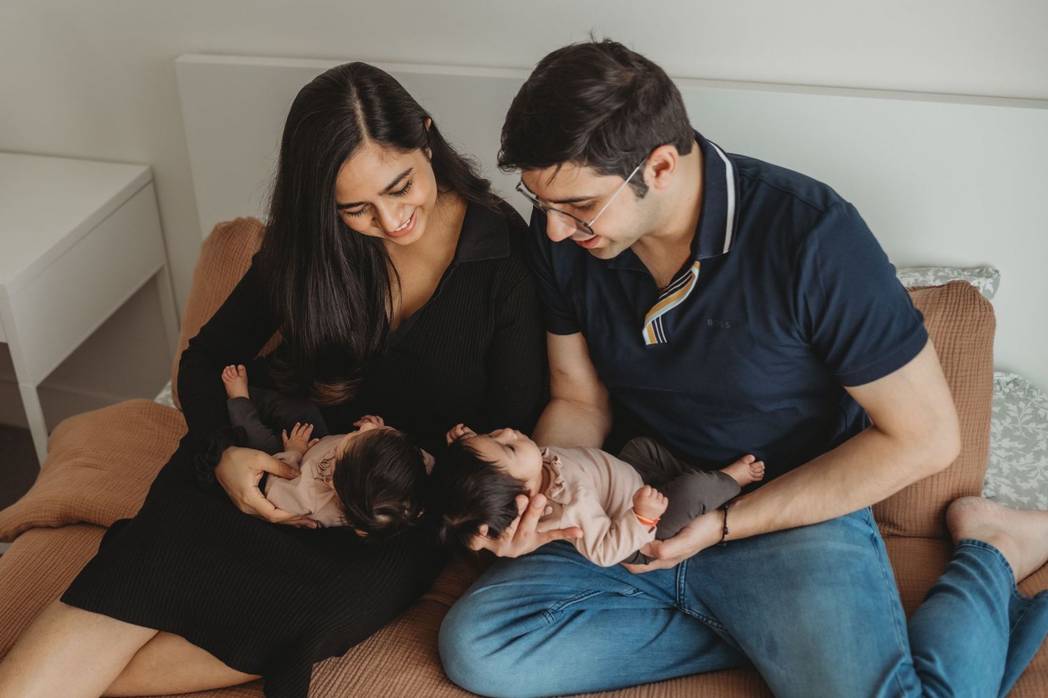 Loving parents sitting together on a couch, each cradling a newborn twin baby in their arms.