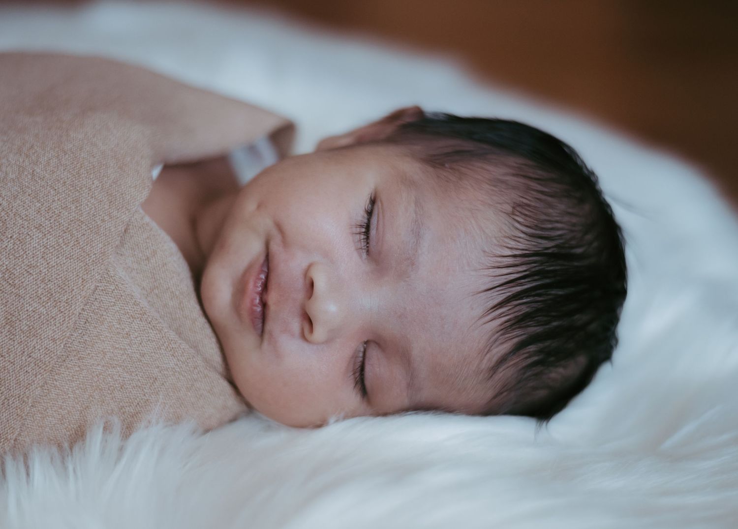 Serene newborn baby smiling while sleeping swaddled in a light pink wrap on a fluffy white blanket.