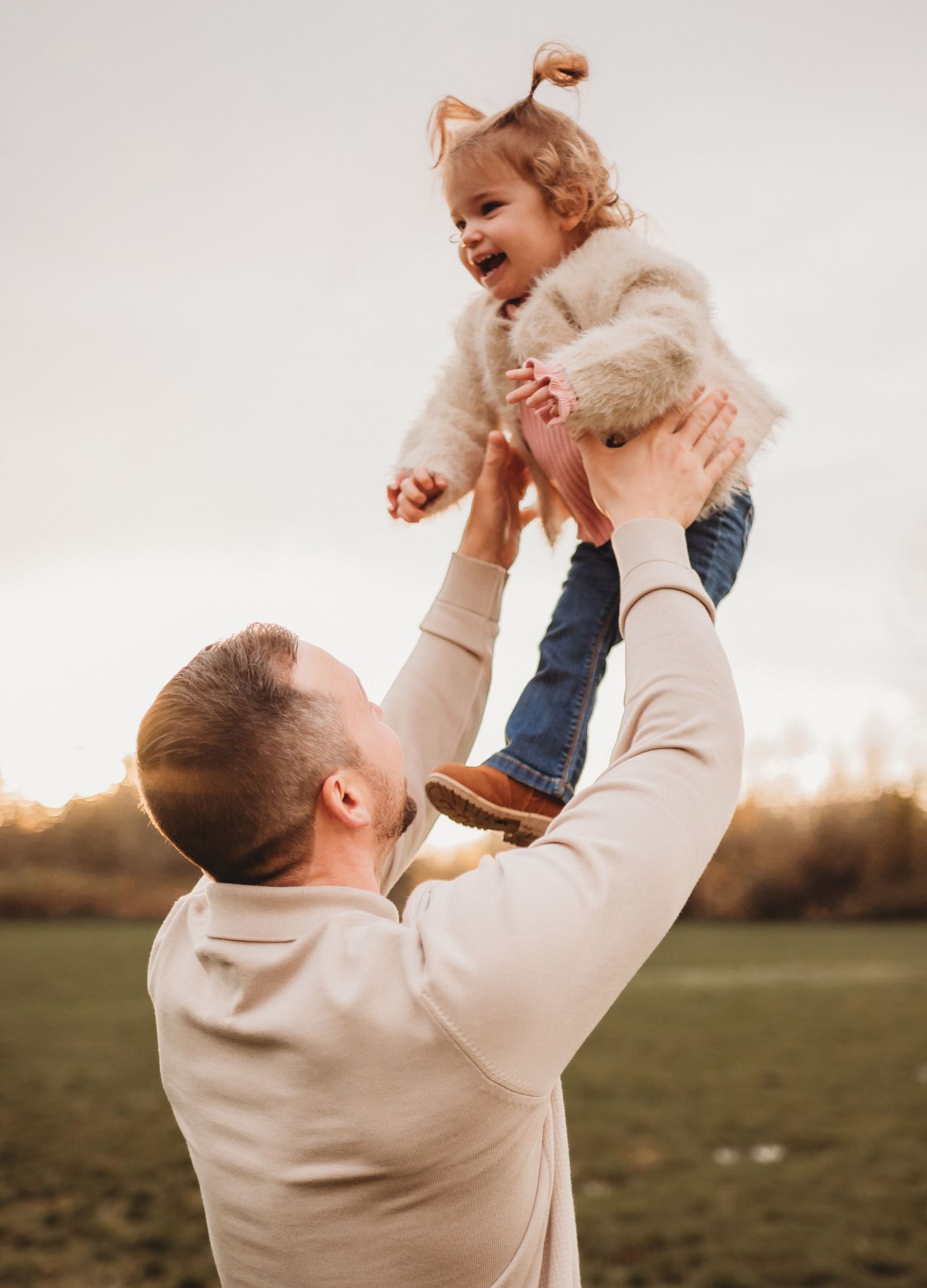 Father joyfully lifting his laughing toddler daughter high into the air outdoors.