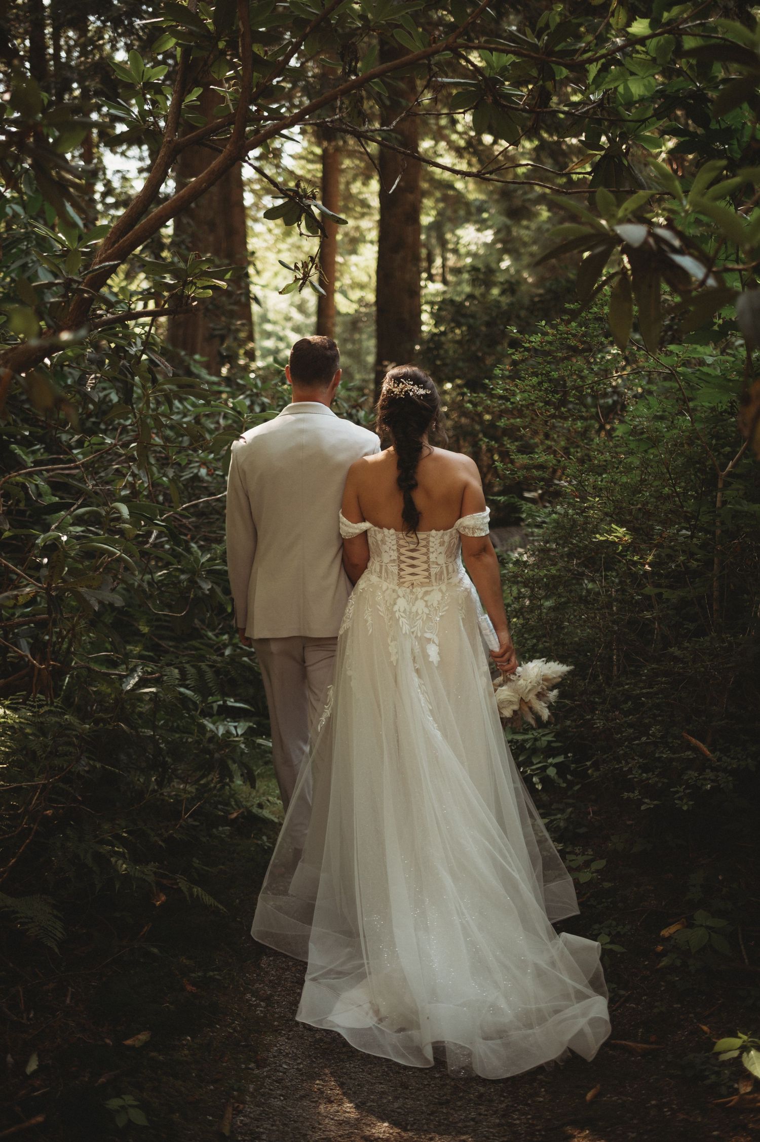 Bride and groom walk hand in hand through a lush green forest path.