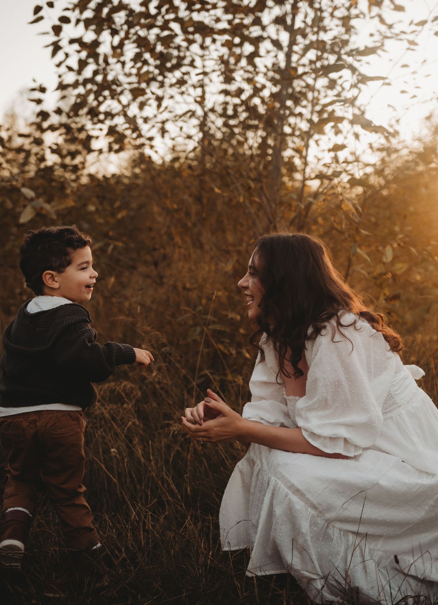 Laughing mother and young son holding hands in a sunlit autumn meadow with golden foliage backdrop.