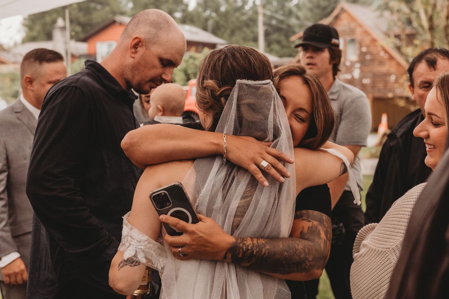 Bride in veil shares emotional embrace with tattooed guest at outdoor wedding reception.