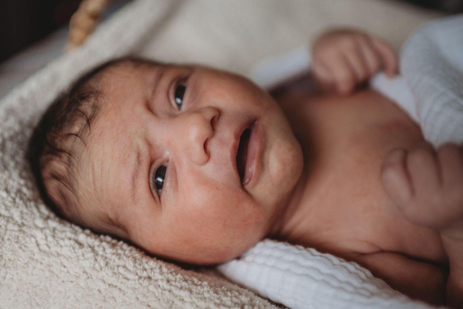 Newborn baby with wide eyes lying on a soft cream textured blanket.