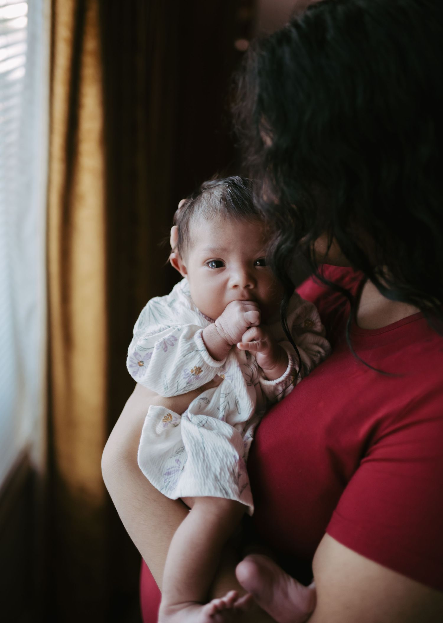 Alert newborn baby in white floral outfit being held by mother in red top.