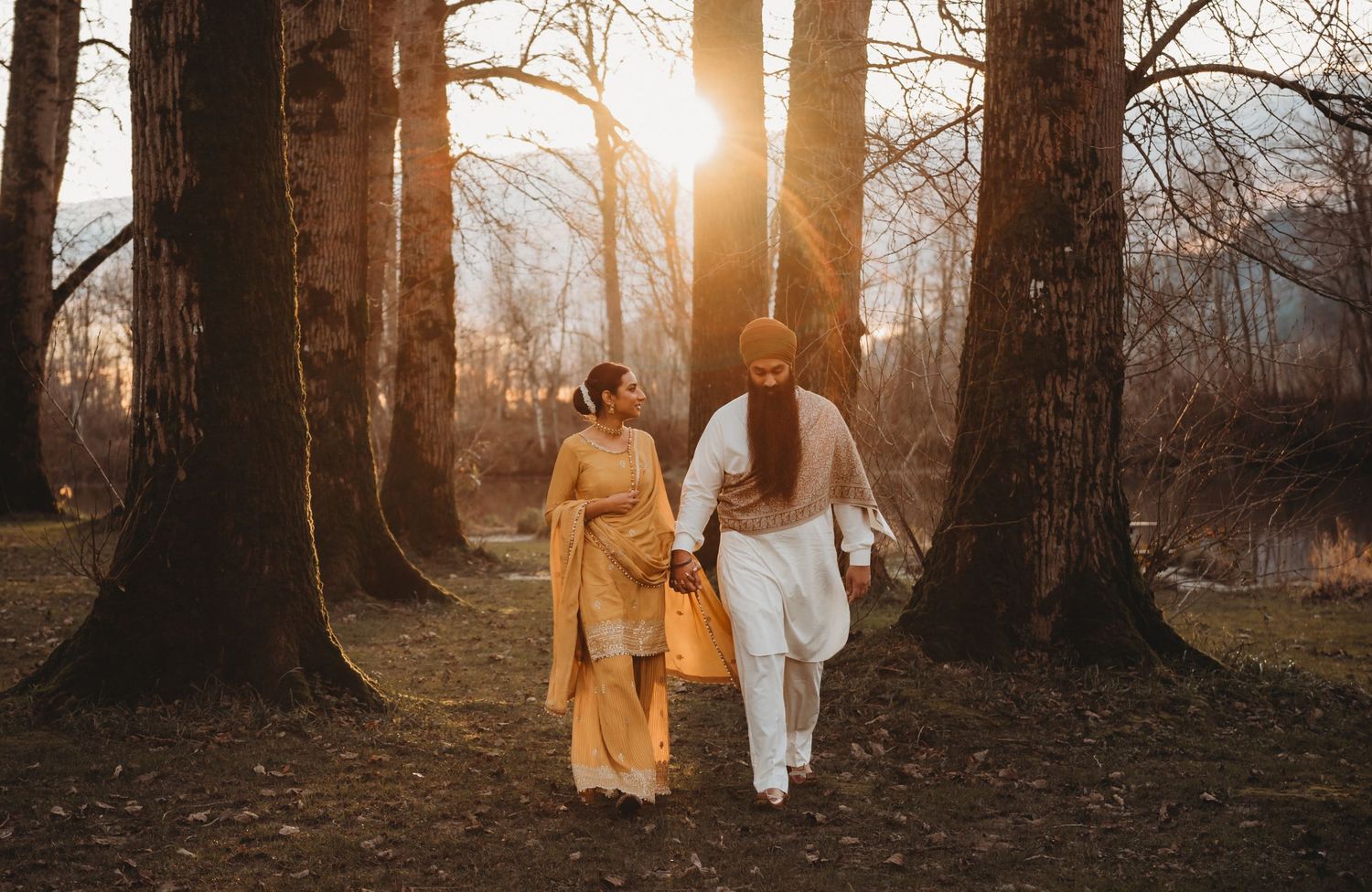 A couple in traditional South Asian clothing walks hand in hand through a forest bathed in golden sunset light.