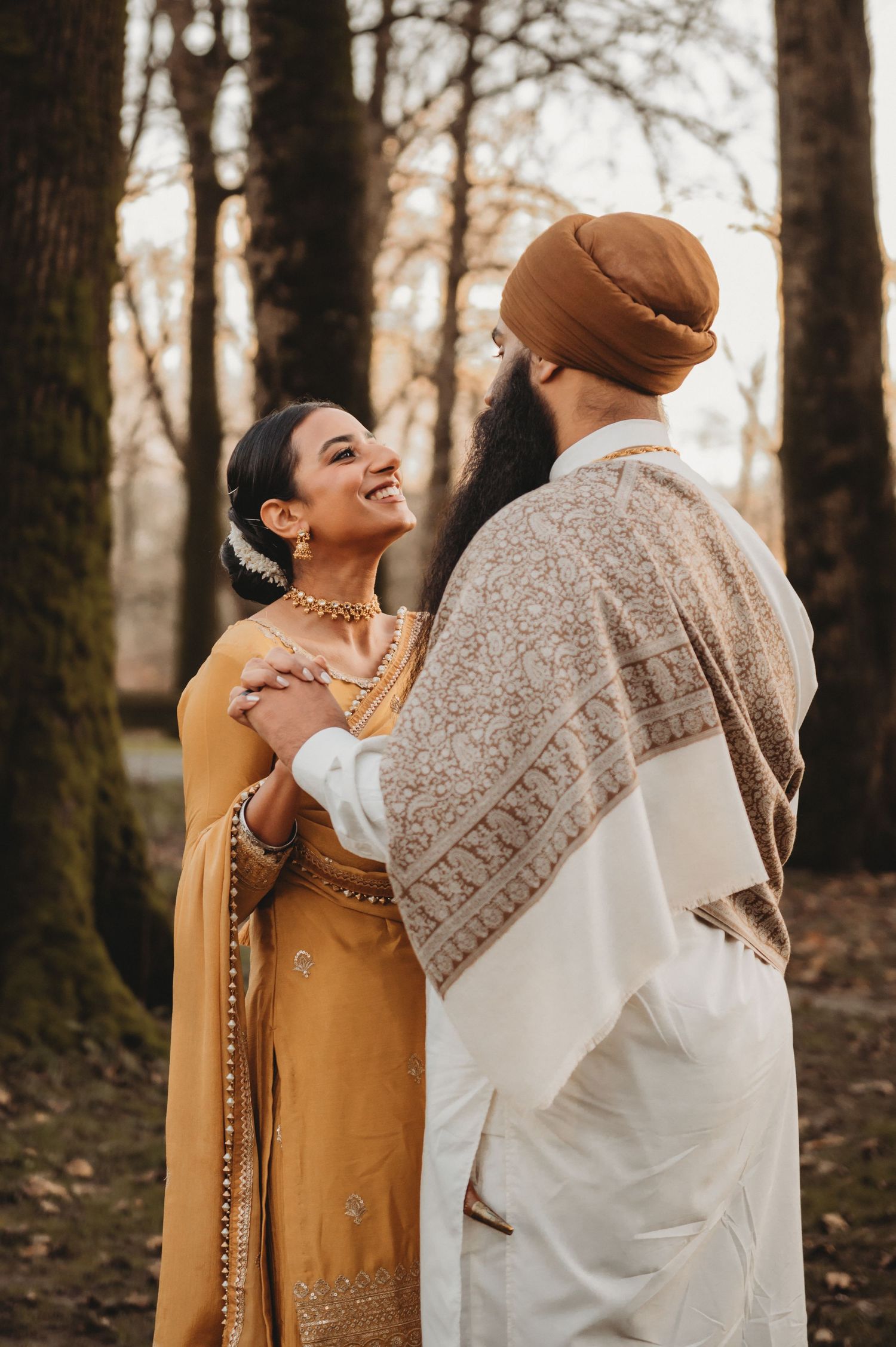 Smiling Sikh bride in mustard salwar embraces groom in embroidered shawl among autumn trees.
