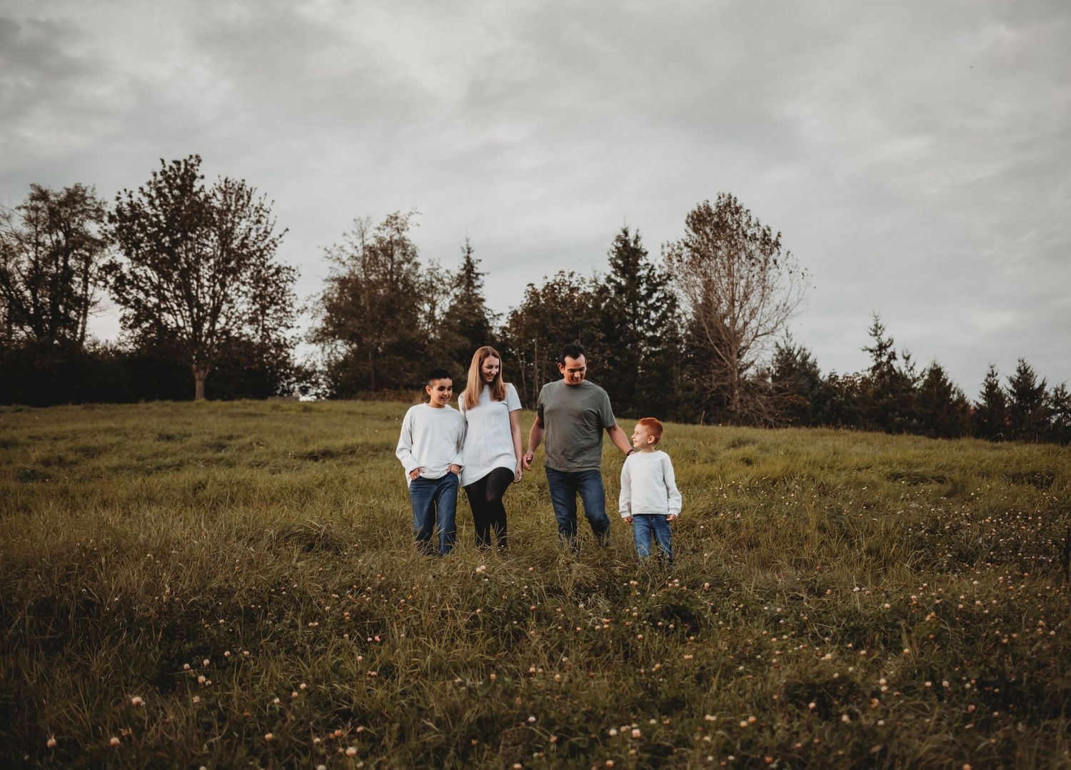 A family of four strolls together across a green meadow with trees in the background.