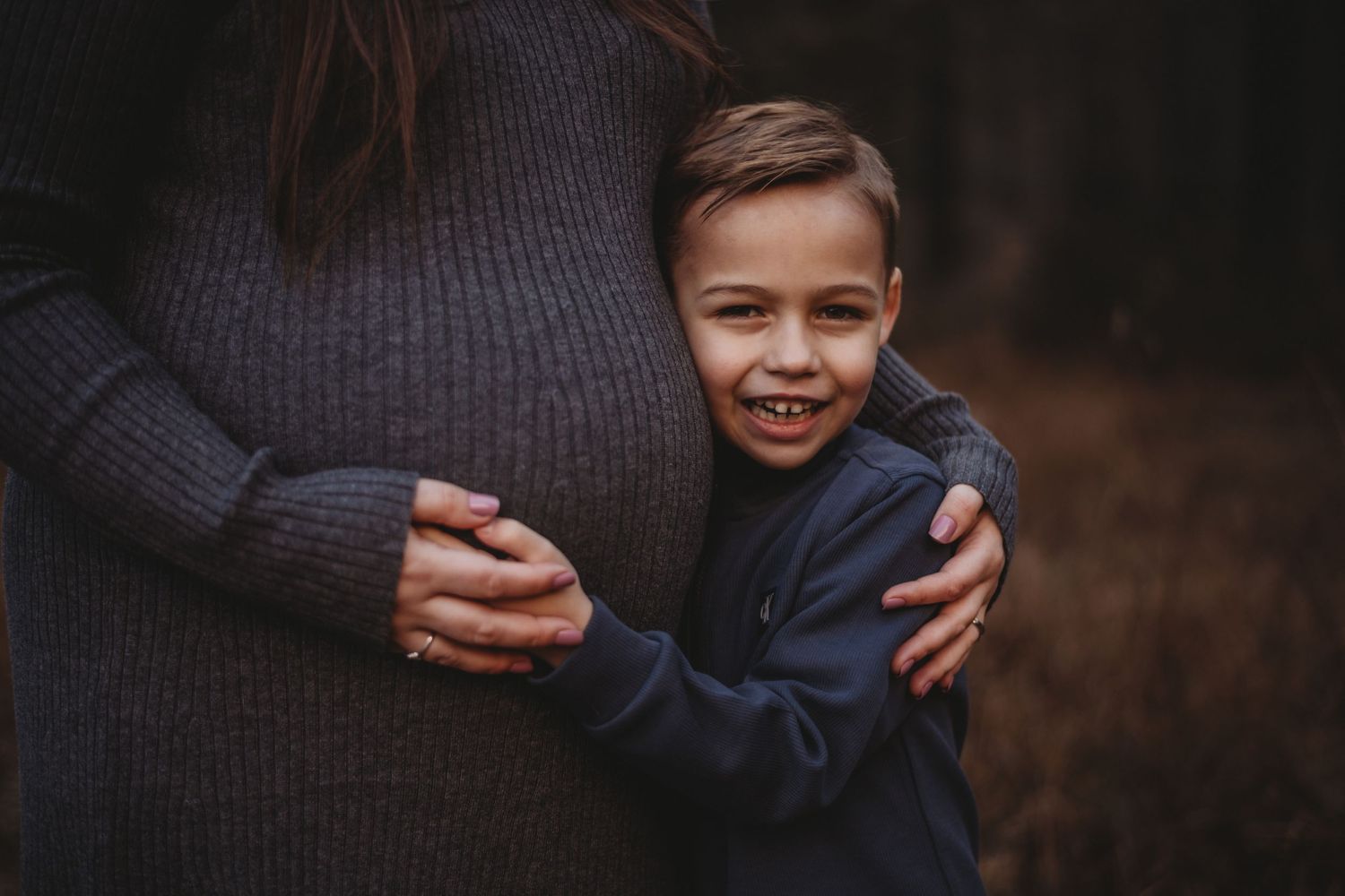 Young smiling boy hugging pregnant mother's belly outdoors in dark wooded setting.
