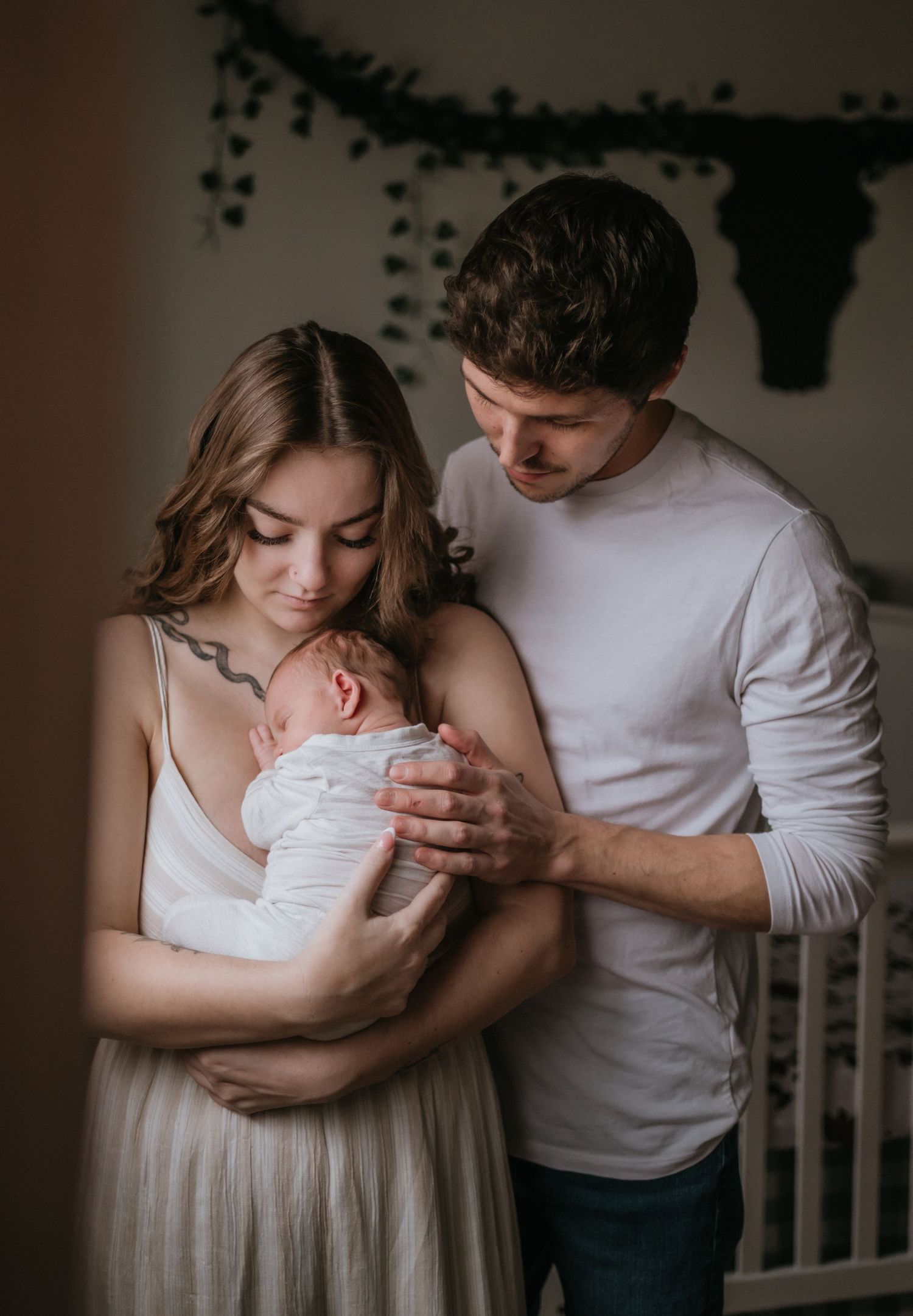 Young couple in white and beige holding swaddled newborn baby in softly lit nursery room.