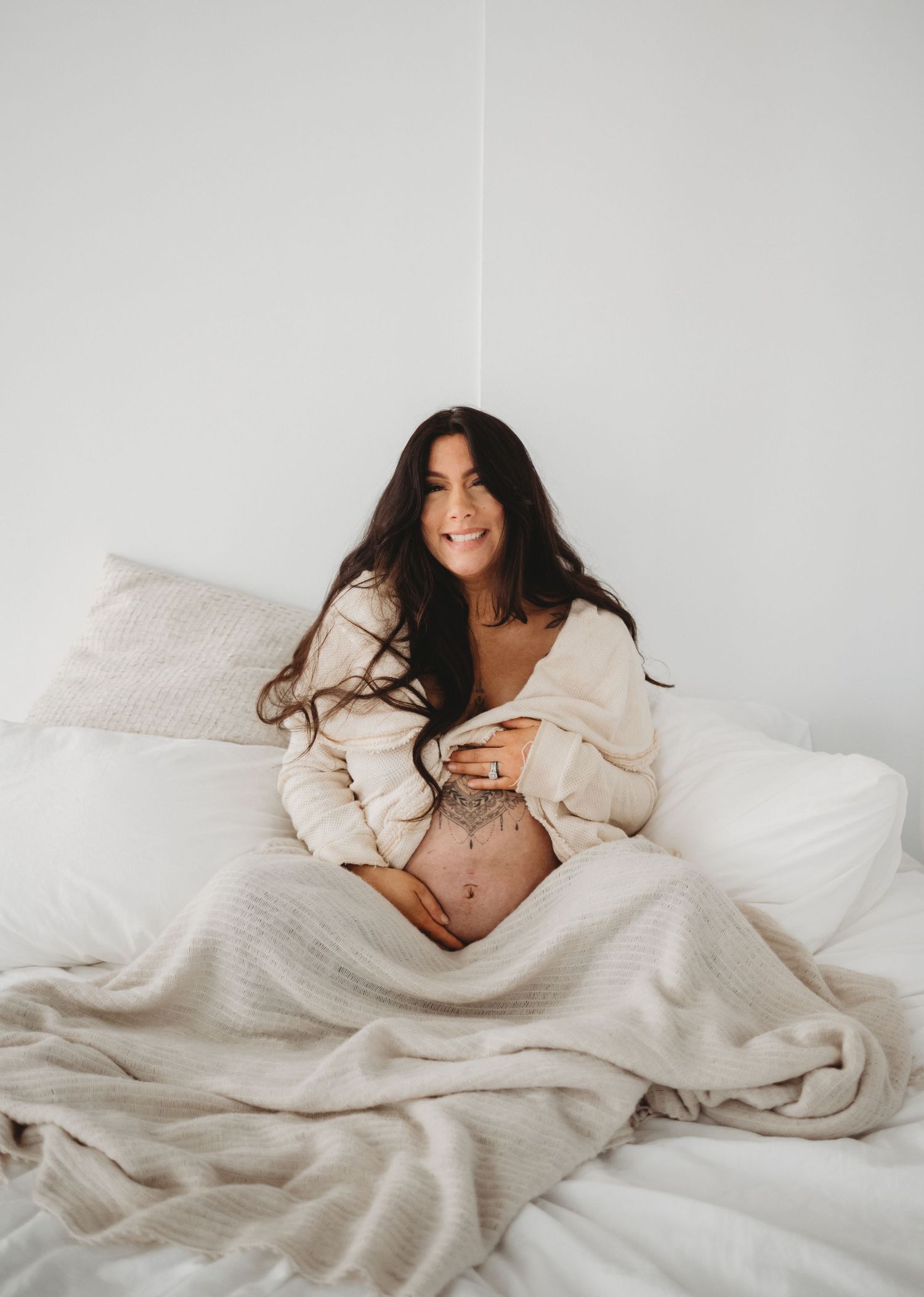 A smiling pregnant woman with tattoos sits on a white bed, cradling her baby bump in a cozy sweater.