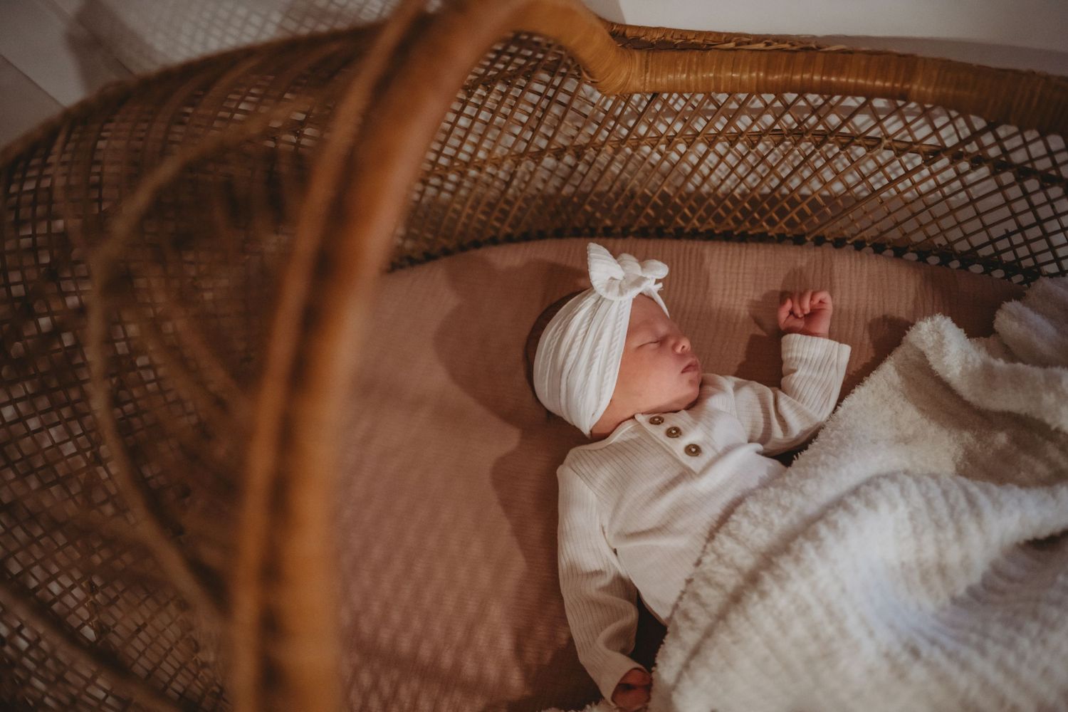 A peaceful newborn baby in a white outfit and bow headband sleeps in a wicker bassinet with a soft blanket.