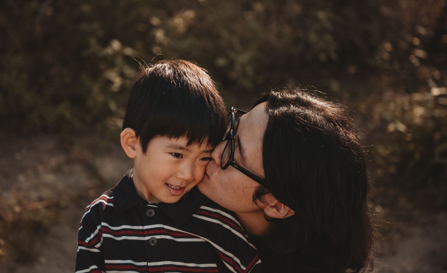 A mother kisses her young smiling boy on the cheek outdoors in warm natural light.