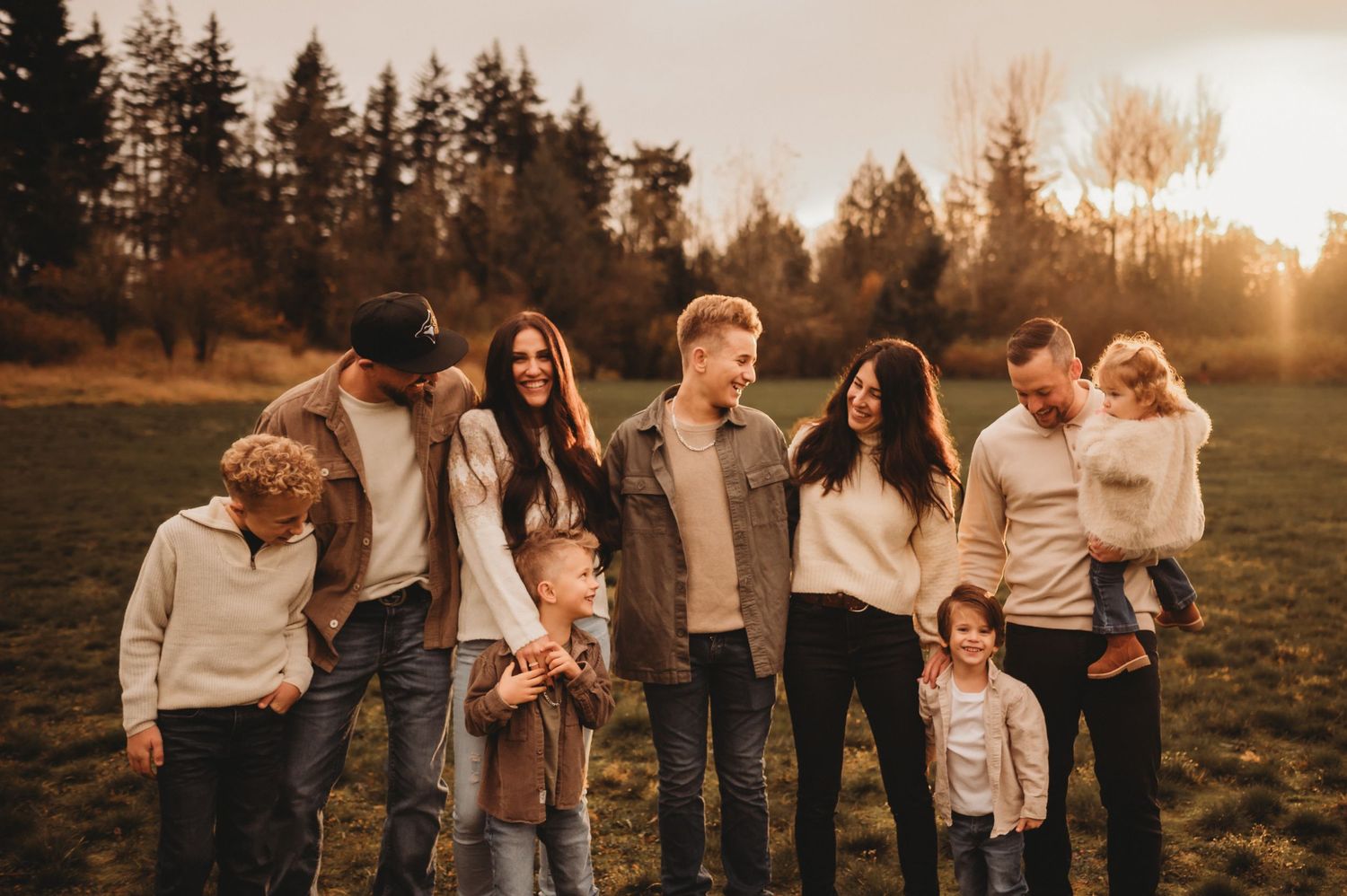 Large family group smiling together in a golden sunset field surrounded by tall trees.