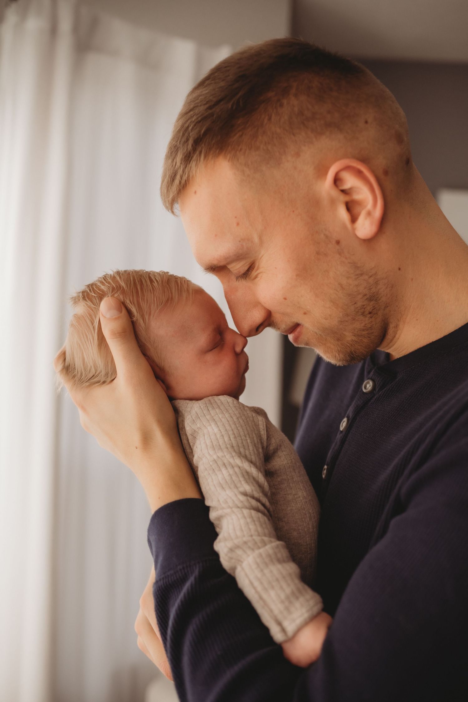 Devoted father holding sleeping newborn baby face-to-face with tender nose-to-nose touch near window.