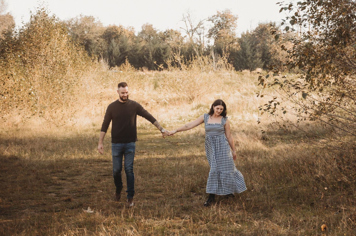 A couple holds hands walking through a sunlit golden meadow surrounded by autumn trees.