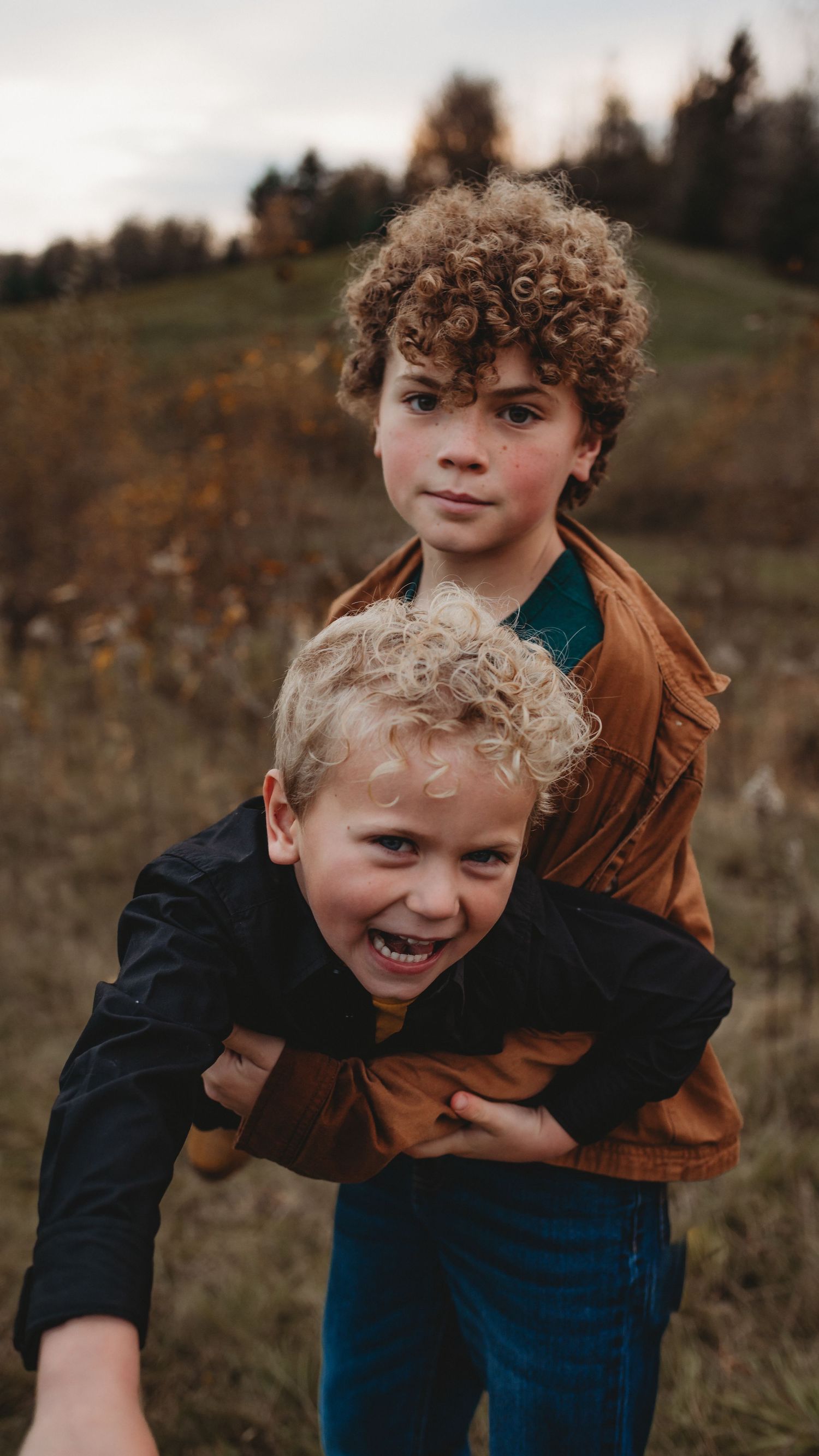 Older curly-haired boy hugging a laughing younger blonde boy in an autumn field.