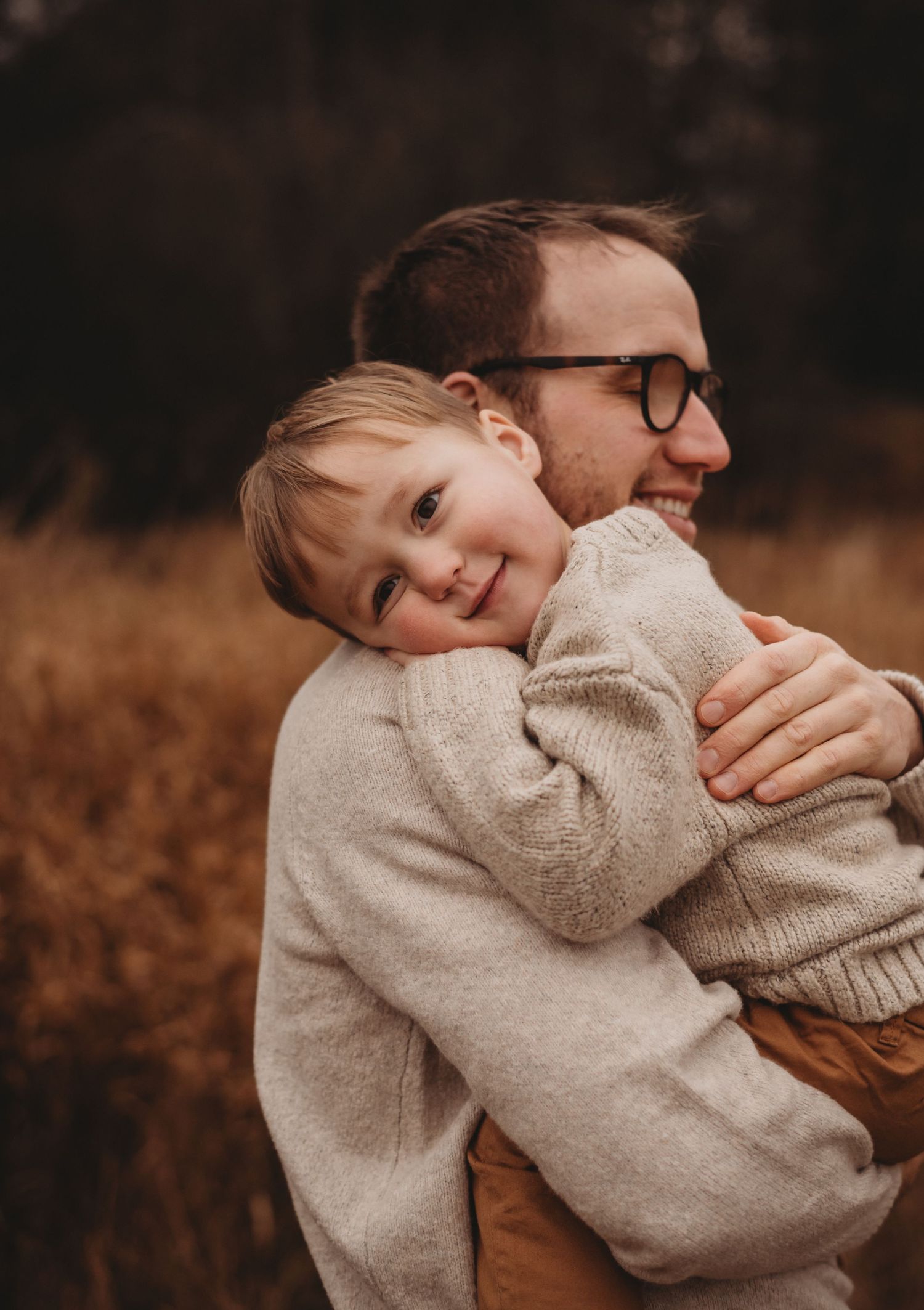 Smiling toddler resting head on fathers shoulder while being carried through autumn meadow.