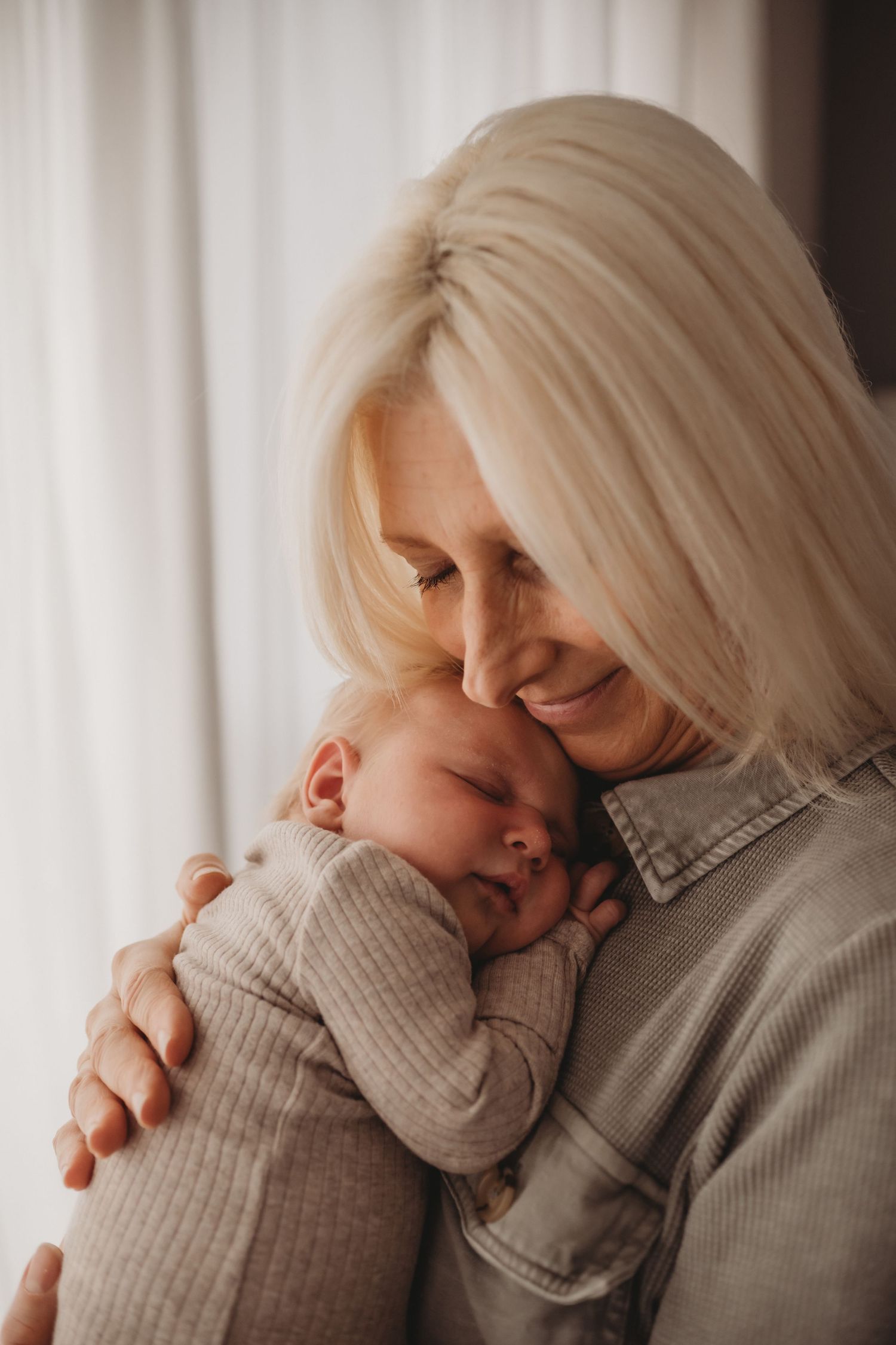 Blonde grandmother smiling warmly while snuggling a peacefully sleeping newborn baby close to her face.