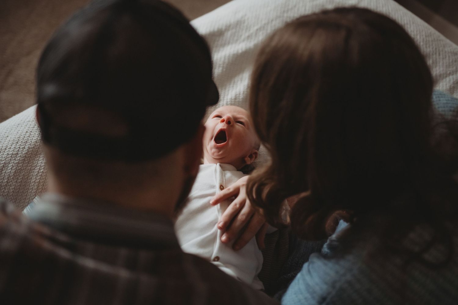 Parents watching their yawning newborn baby resting on a white blanket.