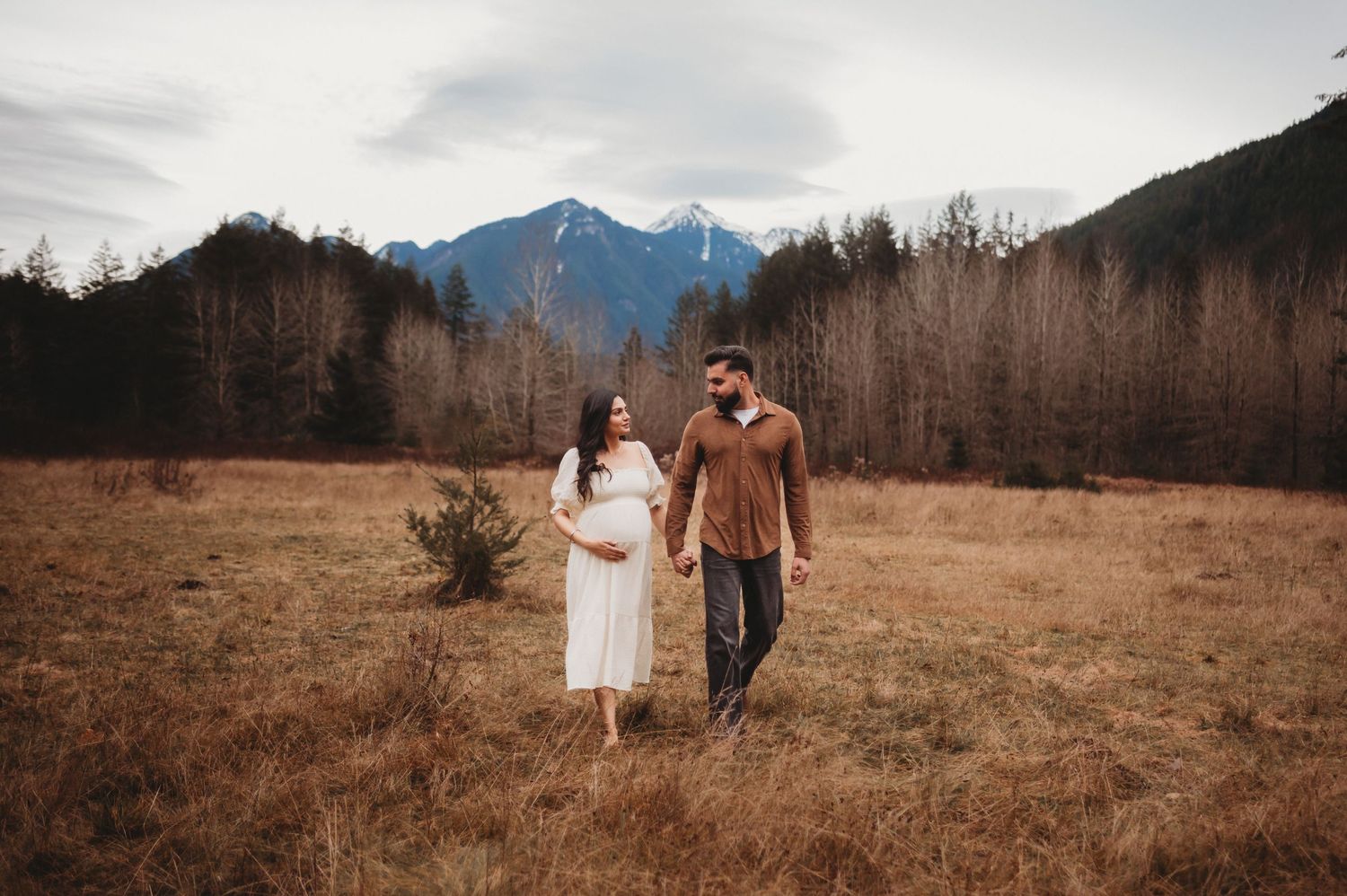 Smiling pregnant couple walking hand-in-hand through golden field with mountain scenery.