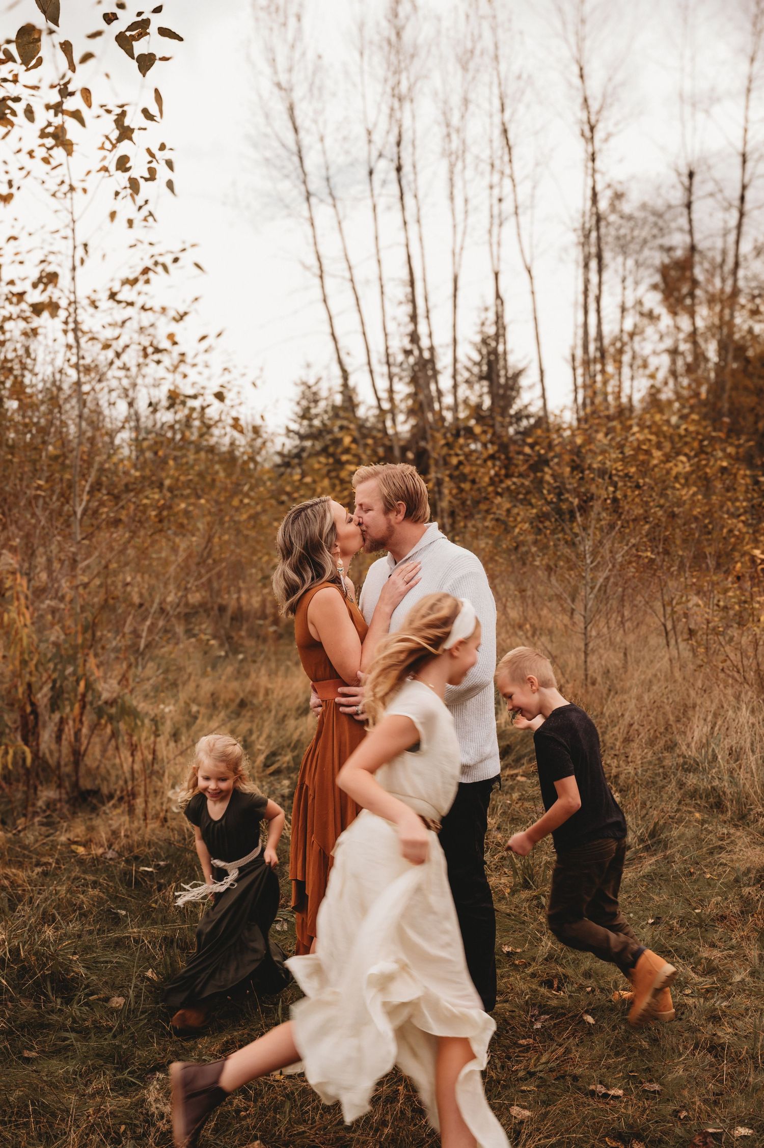 Couple kissing during fall family photo session outdoors while children play around them in golden autumn field.