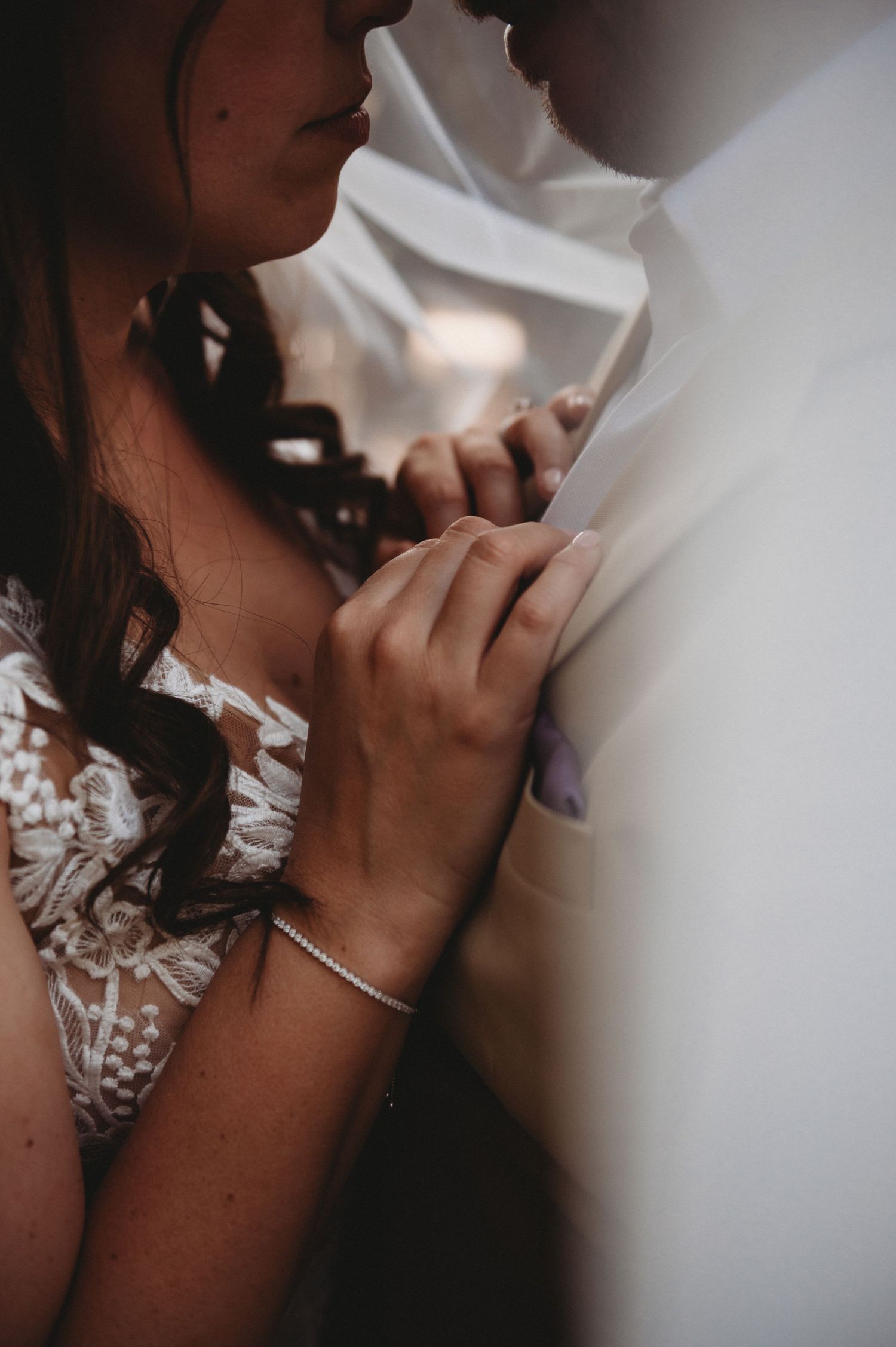 Close-up of bride in lace gown holding groom's shirt during intimate wedding moment.