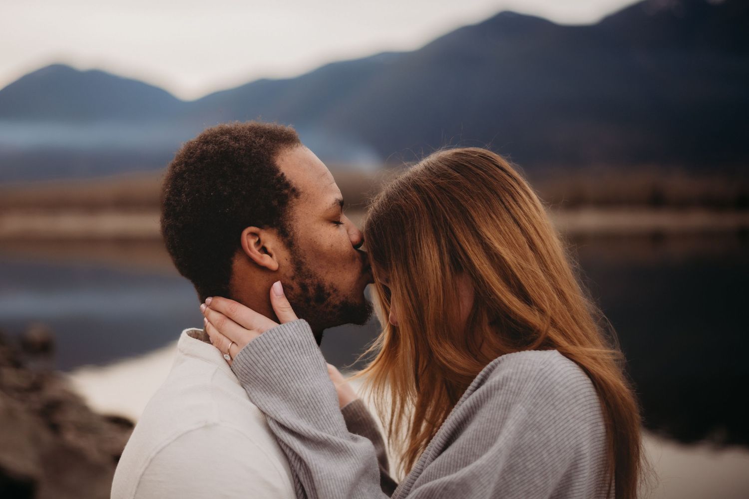 Interracial couple sharing a tender kiss with a scenic mountain lake in the background.