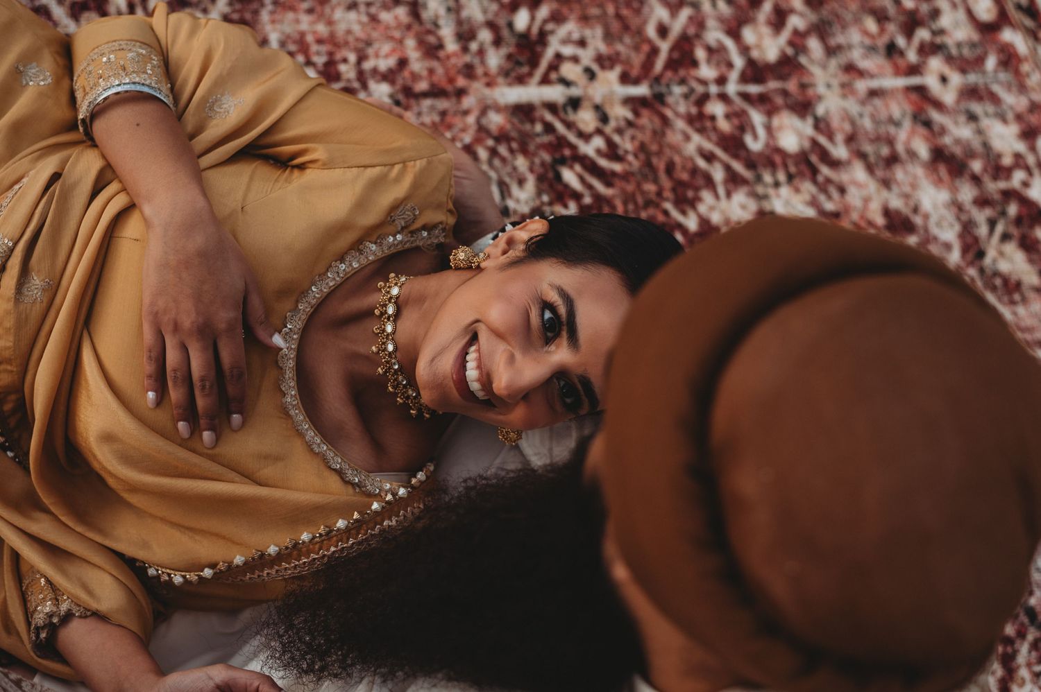 Smiling Sikh bride in gold jewelry lying on a rug looking up at her groom during outdoor photo session.