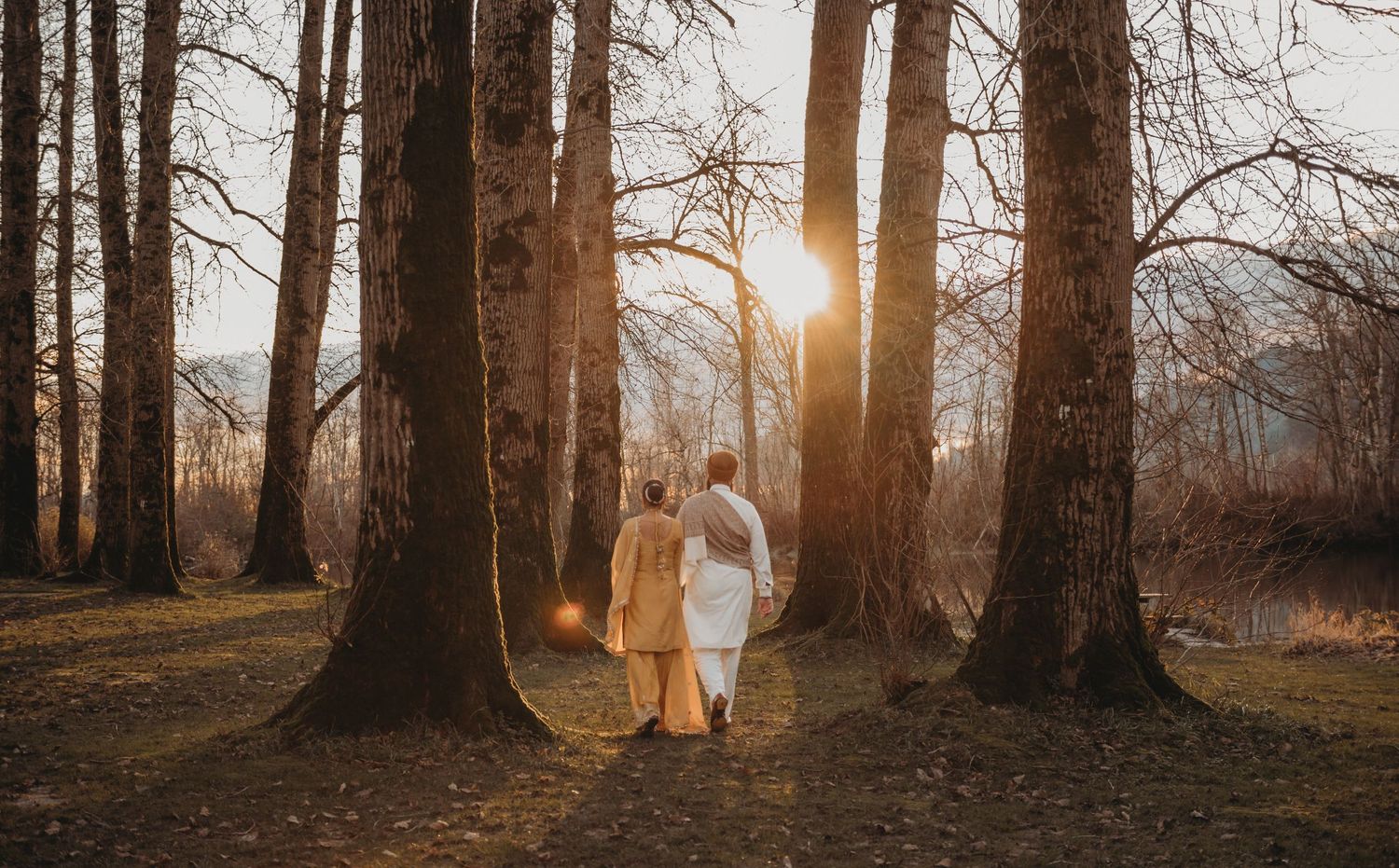 A couple in traditional South Asian attire walks hand in hand through a sunlit forest at golden hour.