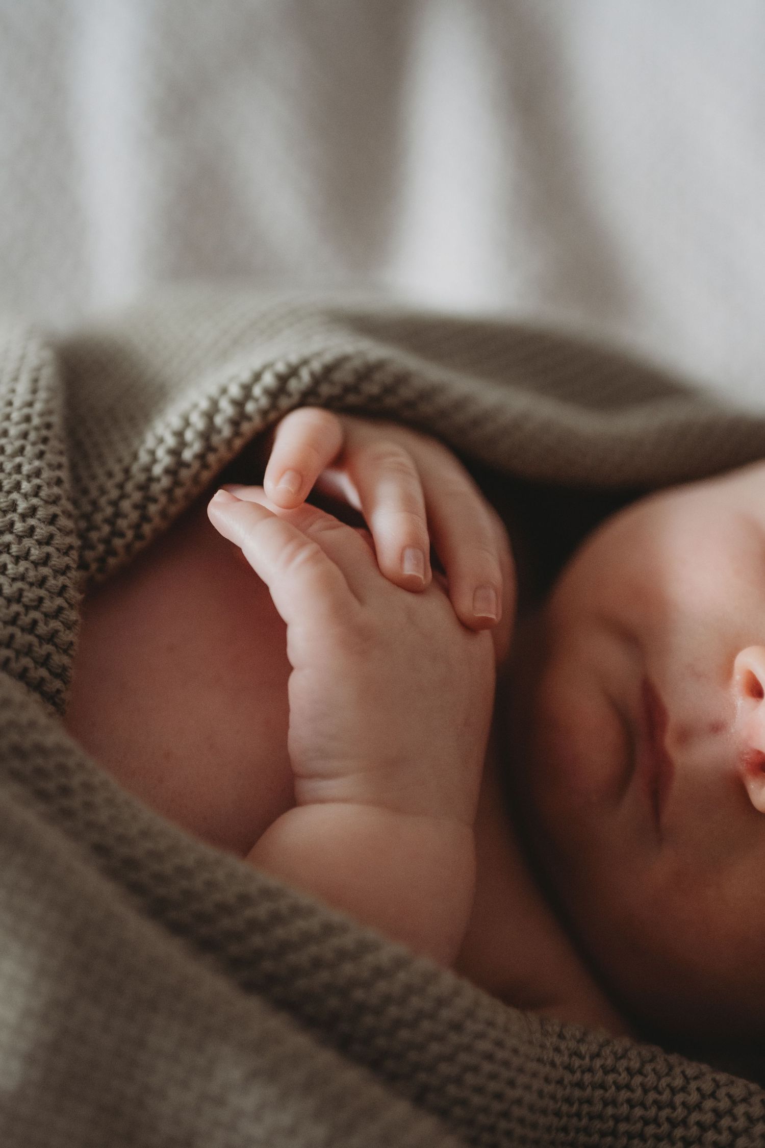 Sleeping newborn baby wrapped in soft grey knit blanket with tiny hands visible, close-up.