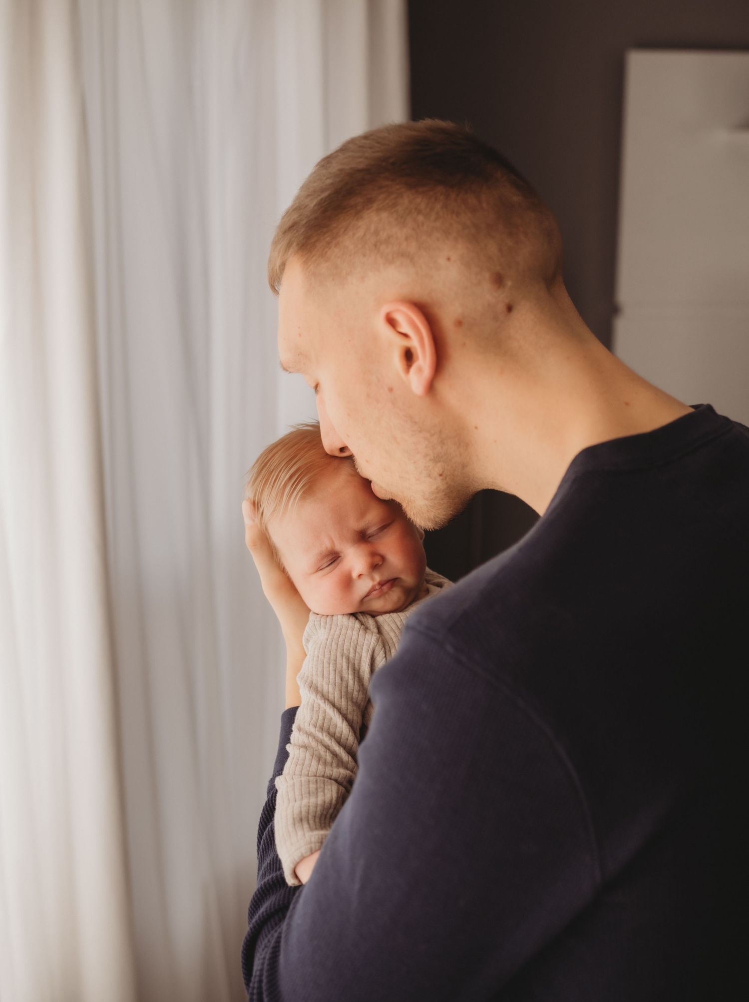 Loving father gently kissing his sleeping newborn baby held close to his chest near window.