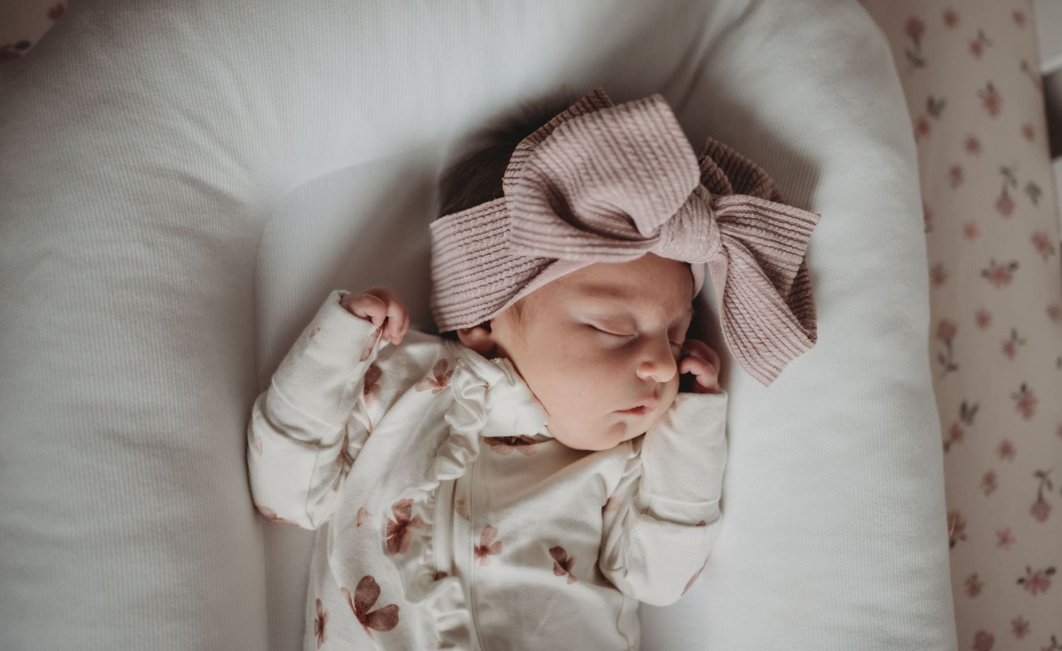 A sleeping newborn baby wearing a floral outfit and pink bow headband rests on white bedding.