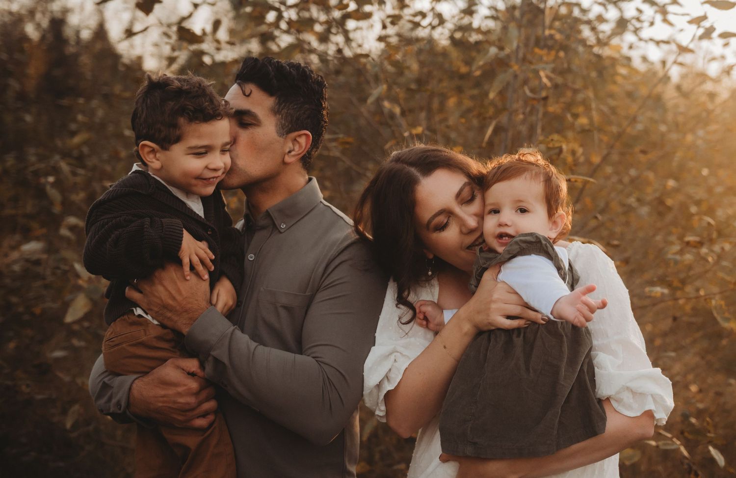 Family of four sharing kisses outdoors in golden autumn light, parents each holding a young child.