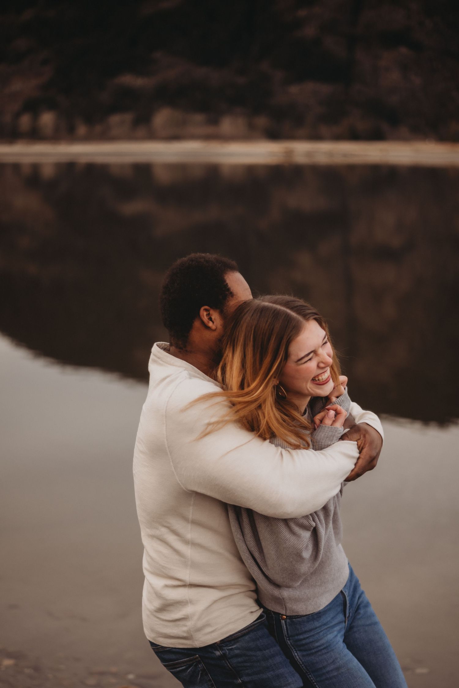 Interracial couple laughing and embracing near a reflective lake outdoors.