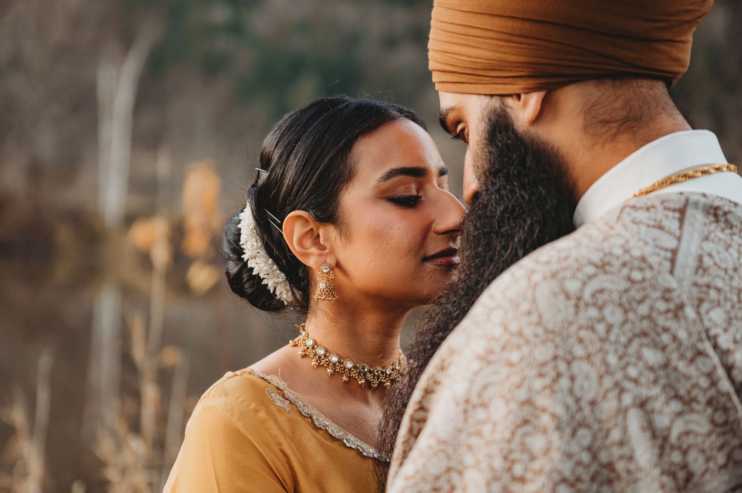 Sikh bride in mustard outfit with gold jewelry leans close to groom in embroidered shawl outdoors.