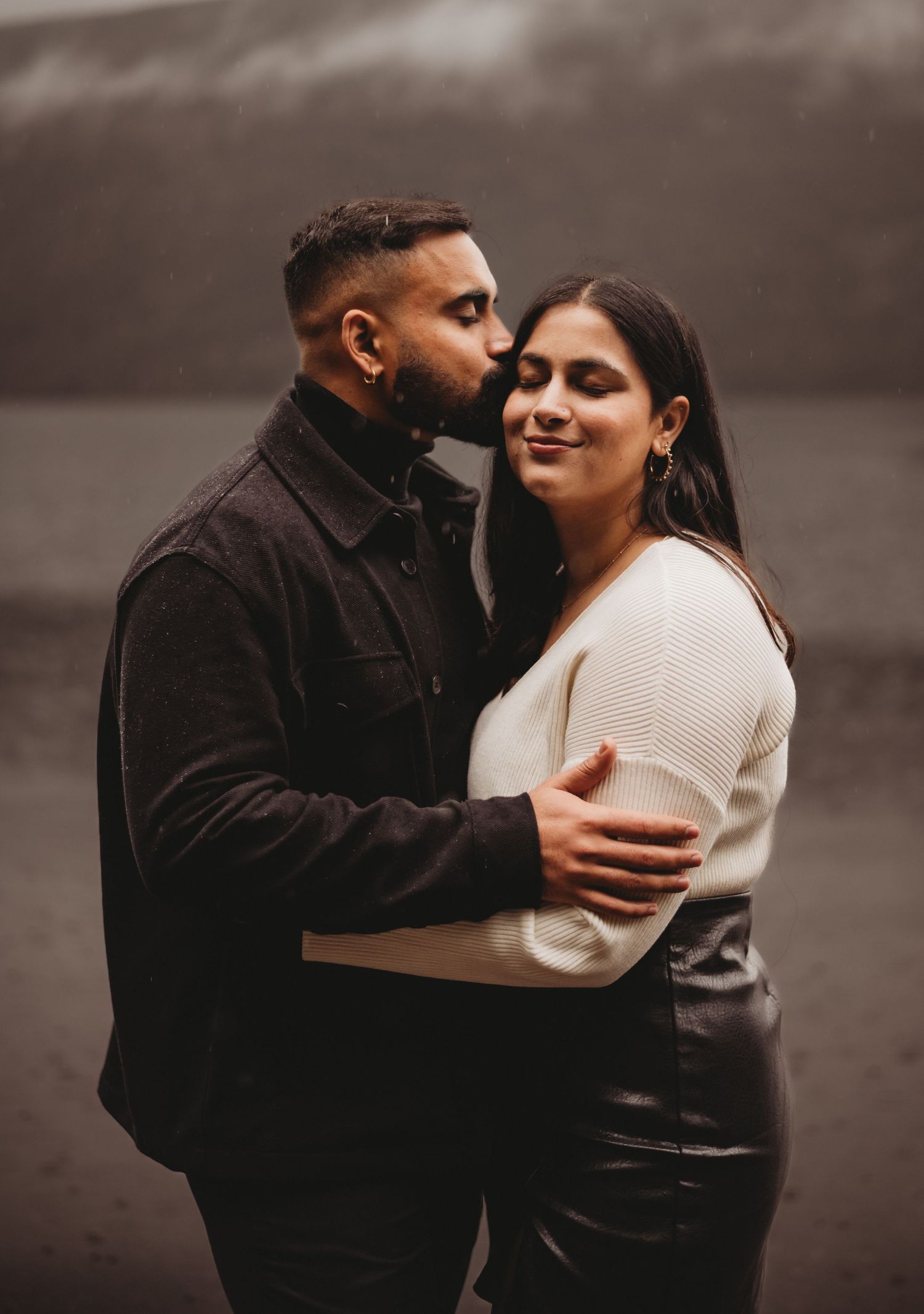Man tenderly kissing woman on cheek while embracing near a foggy lake backdrop.