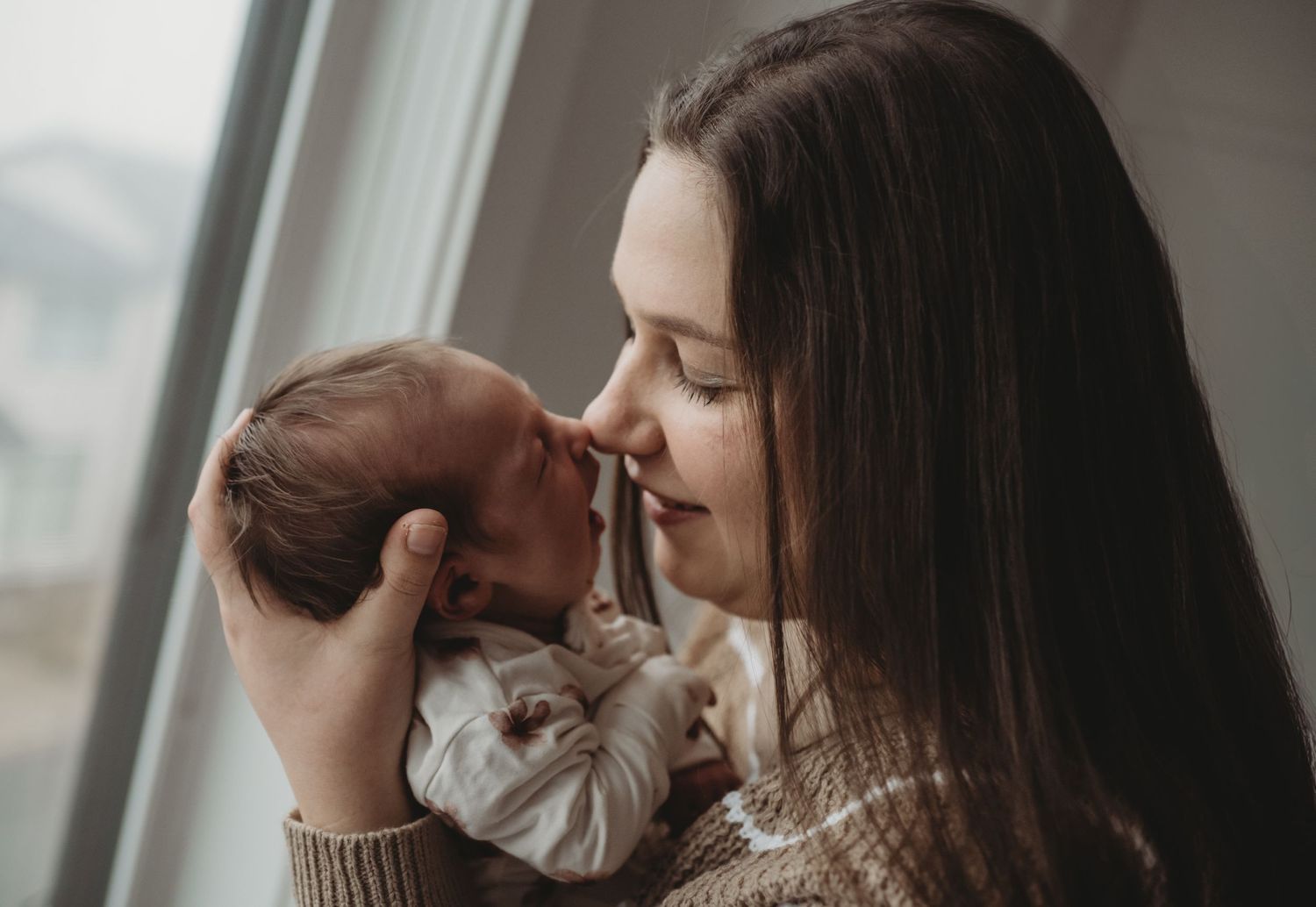 A mother tenderly holds her newborn baby close to her face near a bright window.