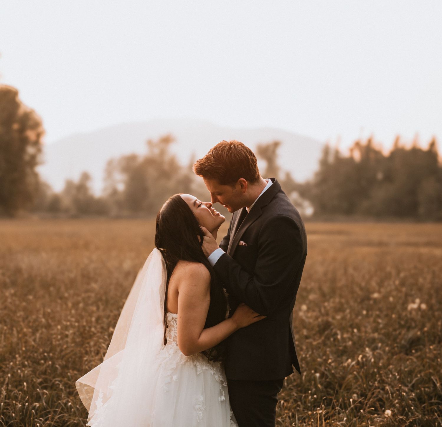 Bride and groom sharing romantic moment in golden field at sunset with mountain backdrop.