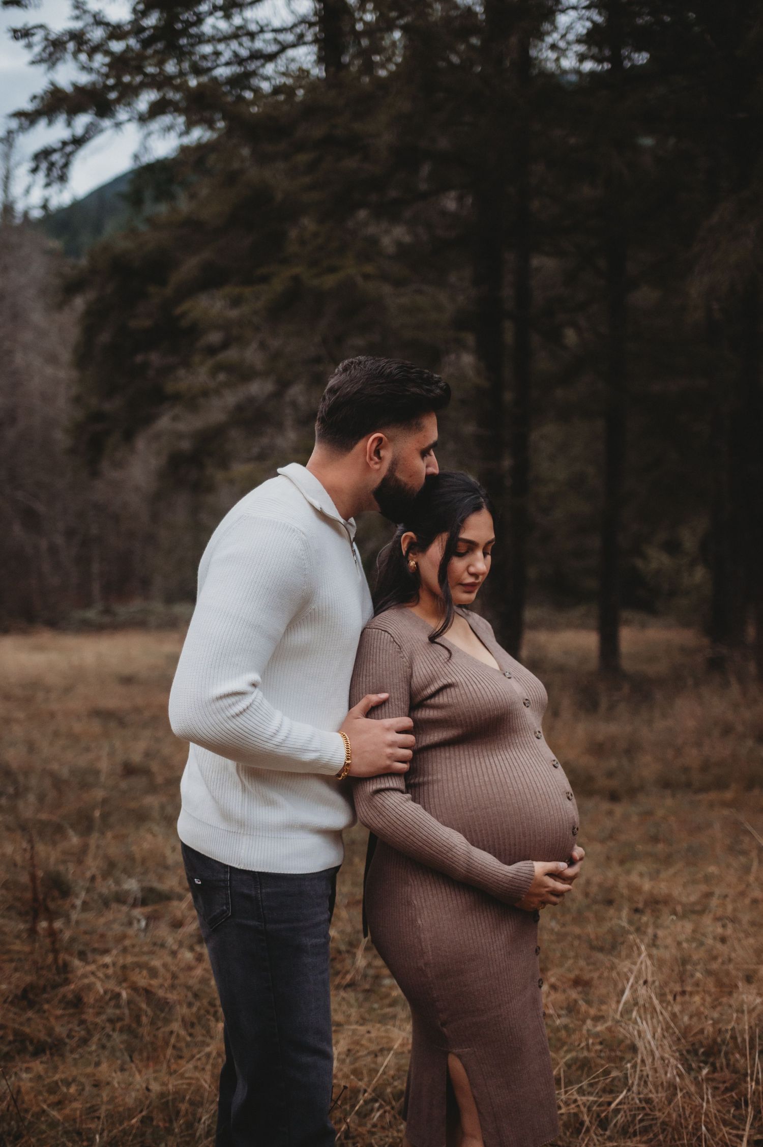 Couple posing together outdoors, man holding pregnant woman's bump in moody woodland maternity photo.