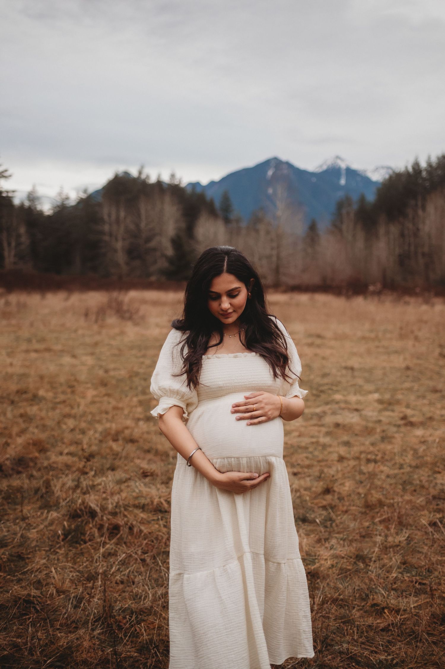 Smiling pregnant woman in white dress gently holds baby bump in scenic outdoor maternity photo.