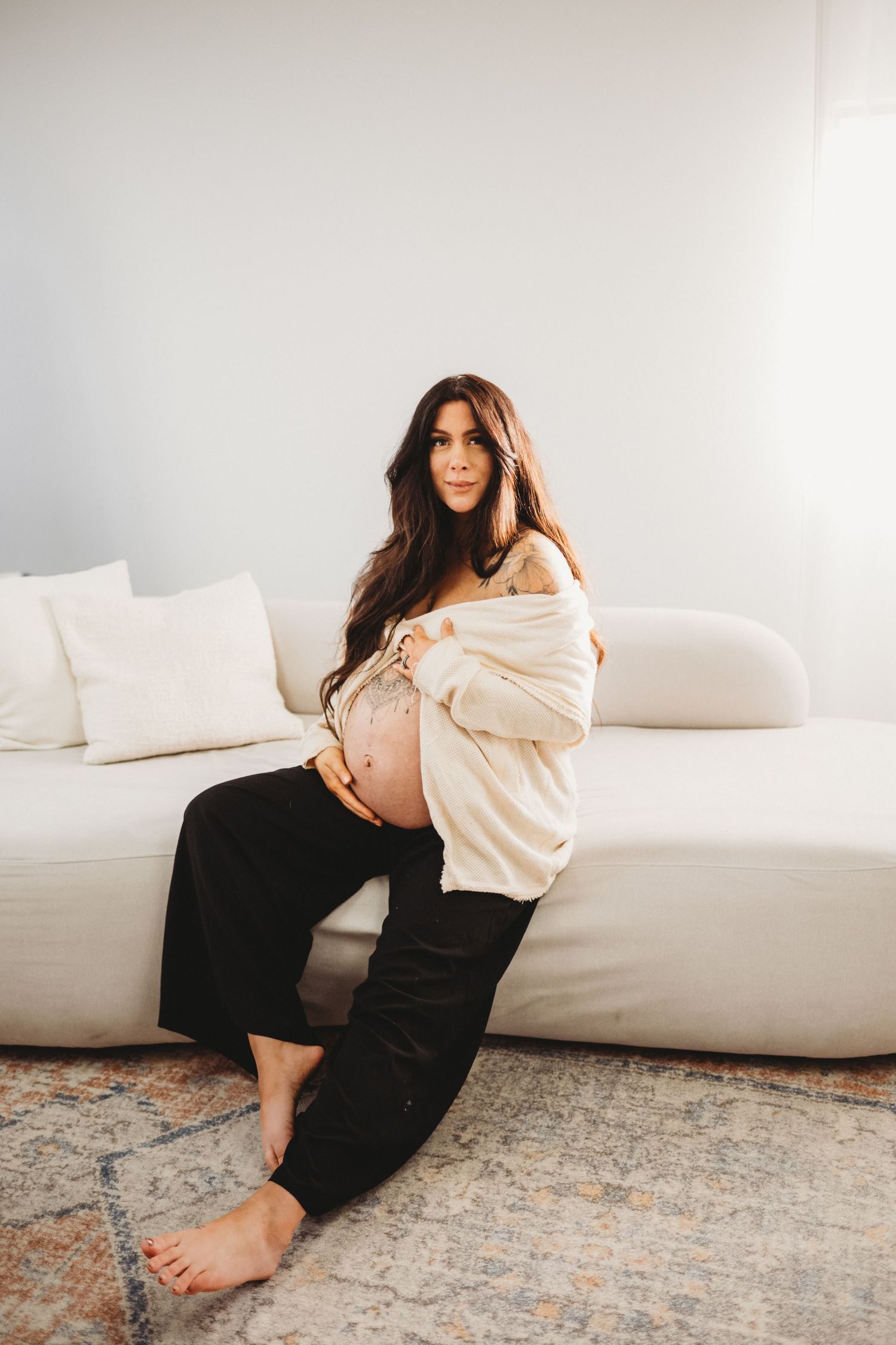 Pregnant woman in cream cardigan and black pants sitting on white sofa cradling baby bump indoors.
