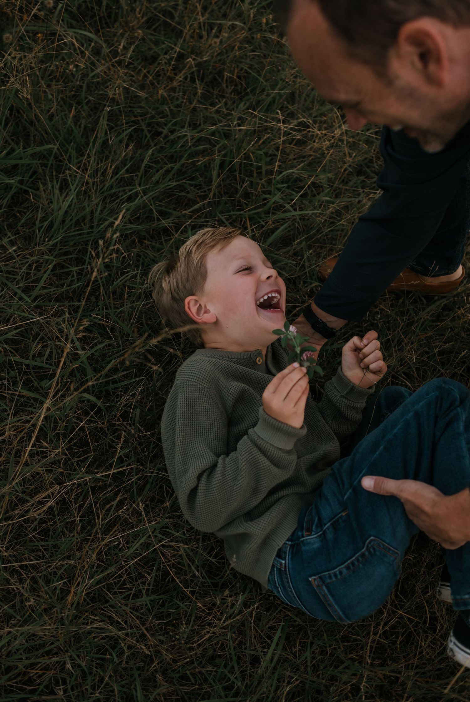 Young boy laughing joyfully while lying in green grass with his father beside him.