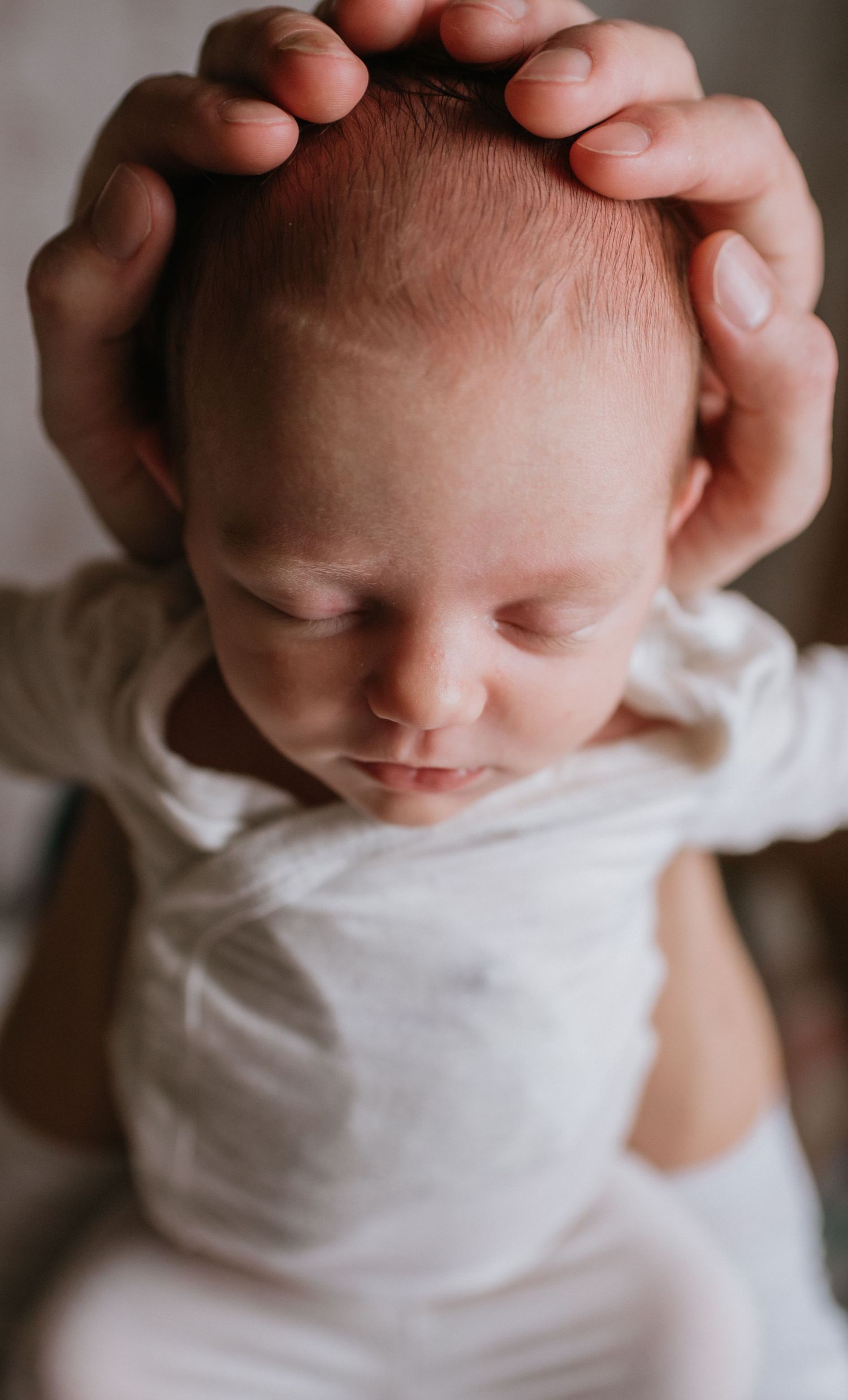 Peaceful sleeping newborn baby in white onesie with parents hands gently resting on head.